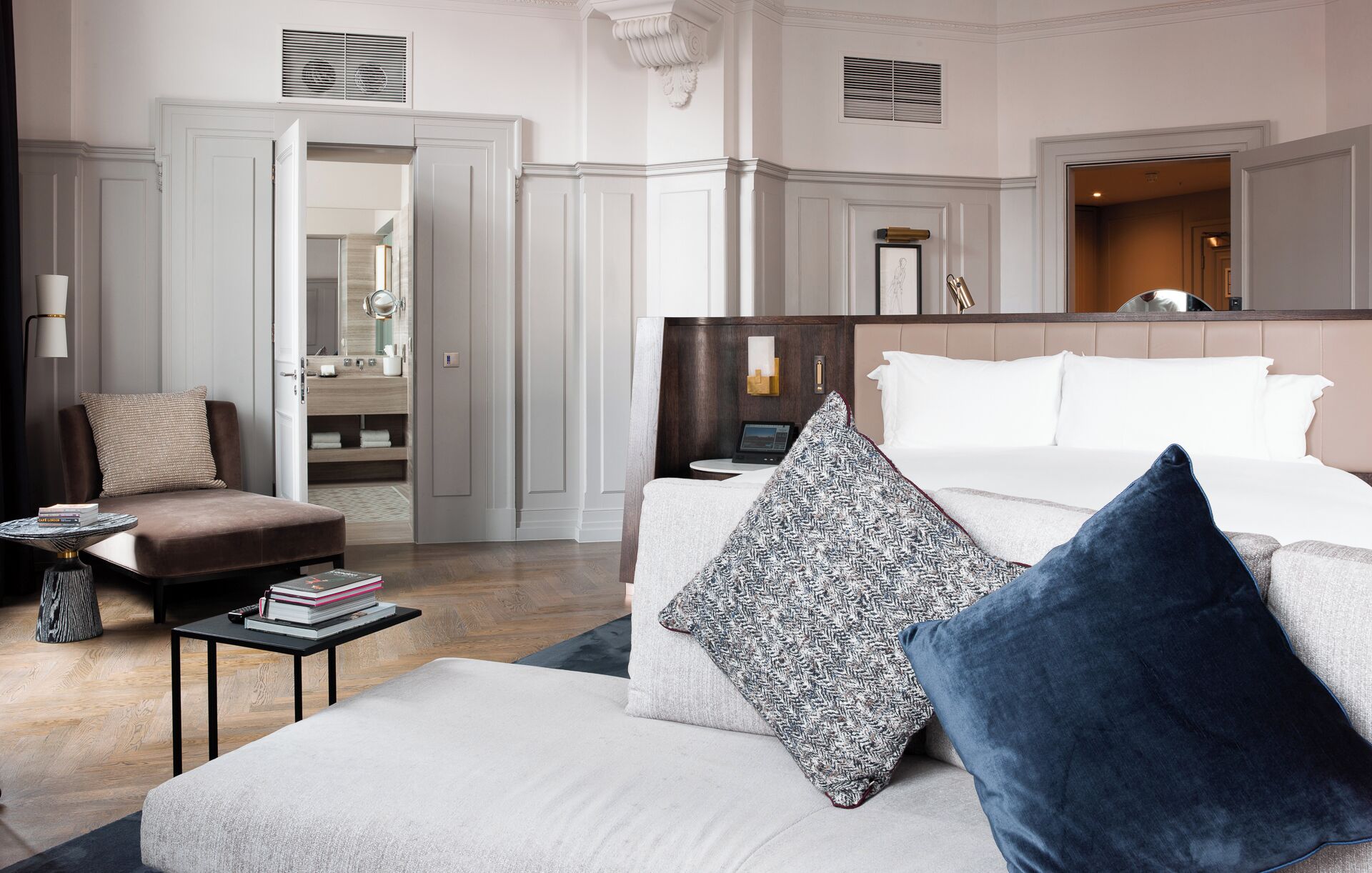 Habitación de hotel con una cama grande, una zona de estar con una silla y una mesita, un baño con espejo y lavabo, y un pasillo con puertas.