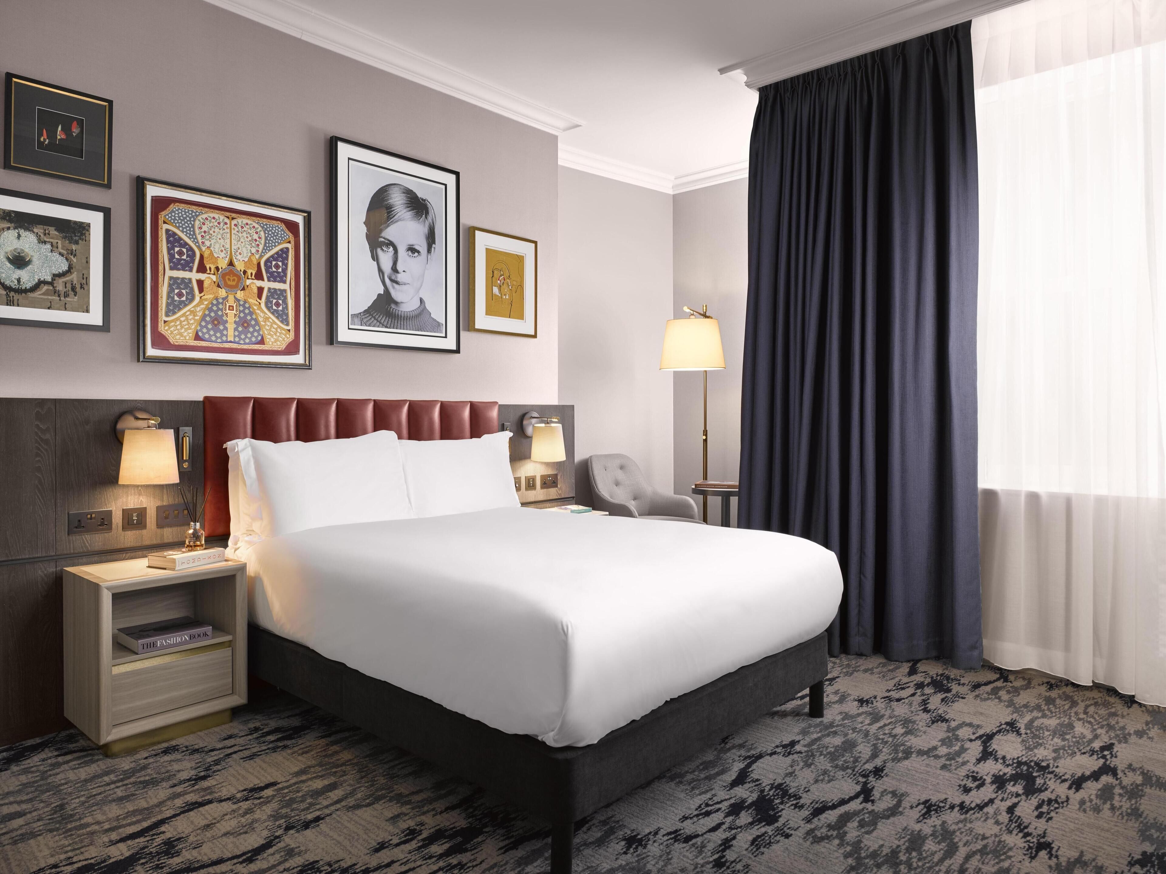 Una habitación de hotel con una cama grande, una mesita de noche con lámpara, una silla y una obra de arte en la pared.
