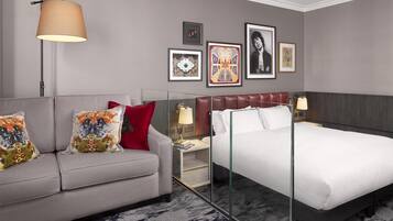 Ein Hotelzimmer mit grauem Sofa, einem Bett mit Glasseitenverkleidung, einer Lampe und gerahmten Kunstwerken an der Wand.