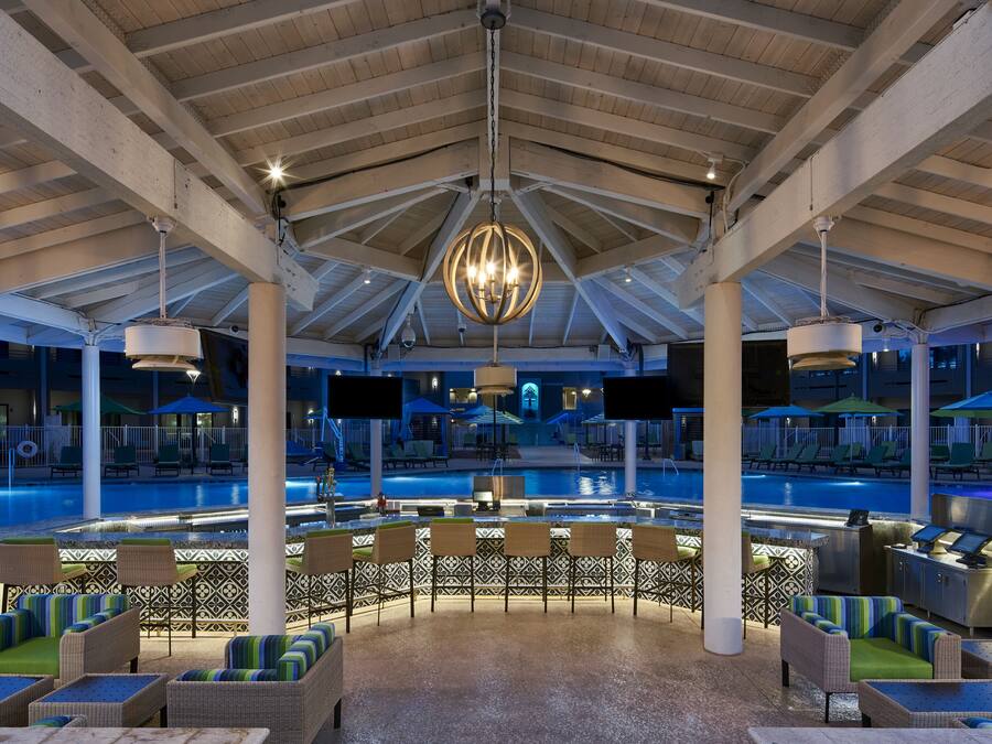A poolside area with lounge chairs and a bar, under a high ceiling with exposed beams.