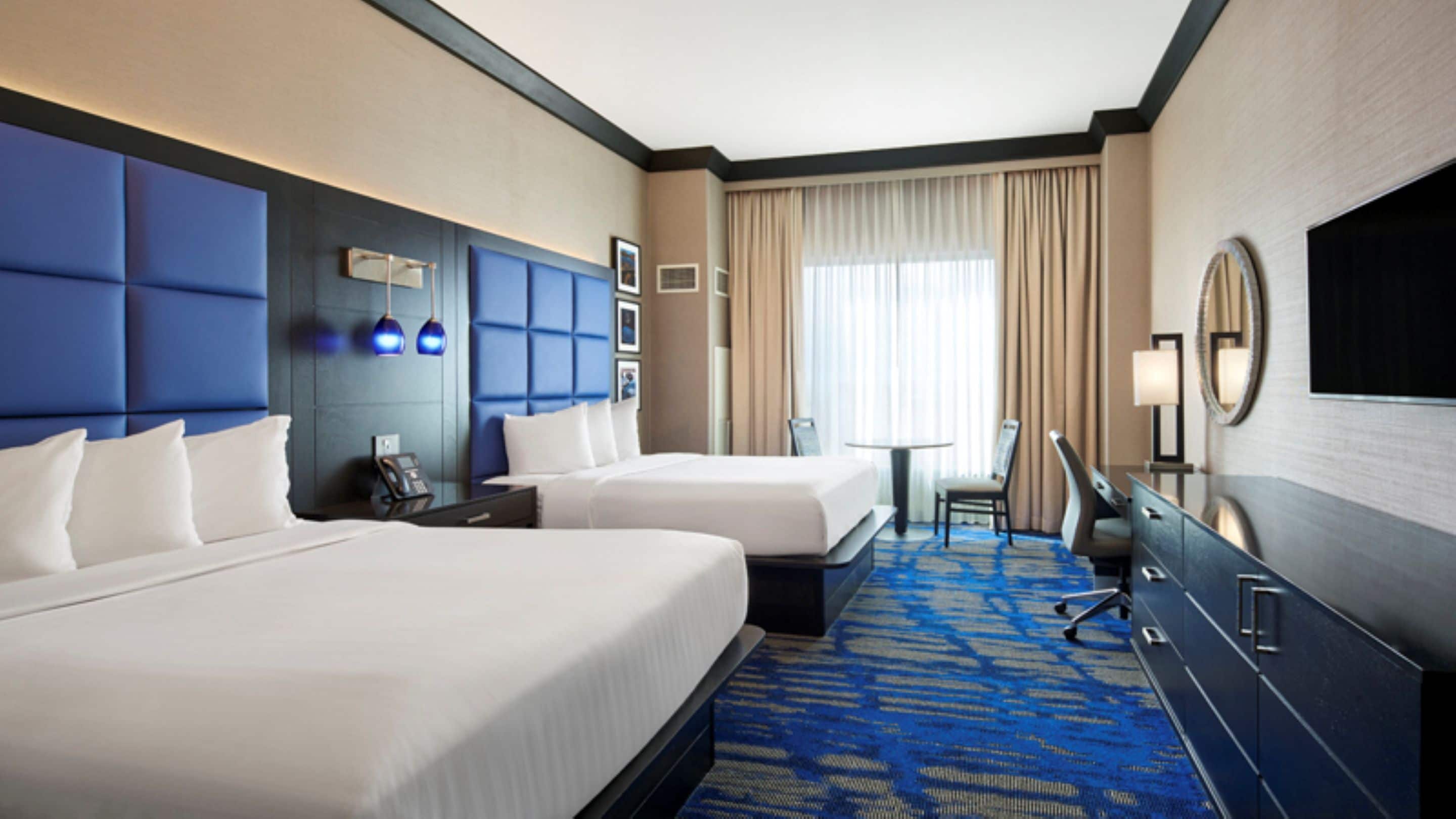 A hotel room with two beds, a desk, a chair, a TV, and a large window with curtains.
