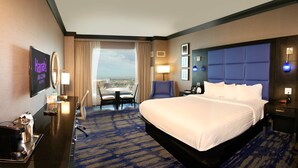 Ein Hotelzimmer mit einem großen Bett, einem Schreibtisch mit Telefon, einem Sessel, einem Fernseher und Blick auf die Stadt.