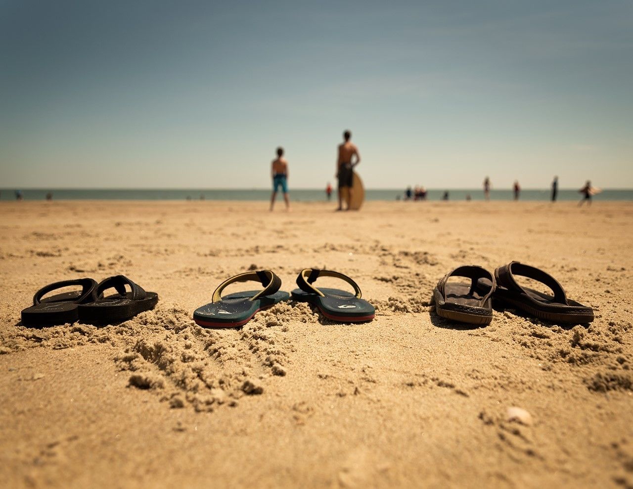 A pair of flip-flops on a sandy beach with people in the background.