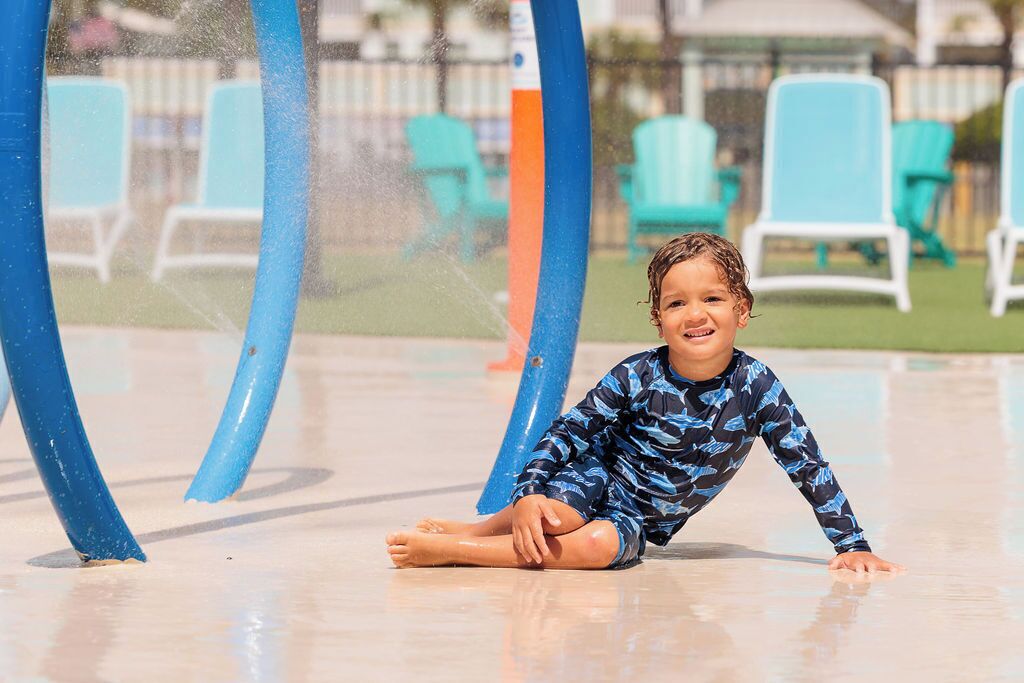 Un niño sentado sobre una superficie mojada, con chorros de agua al fondo.