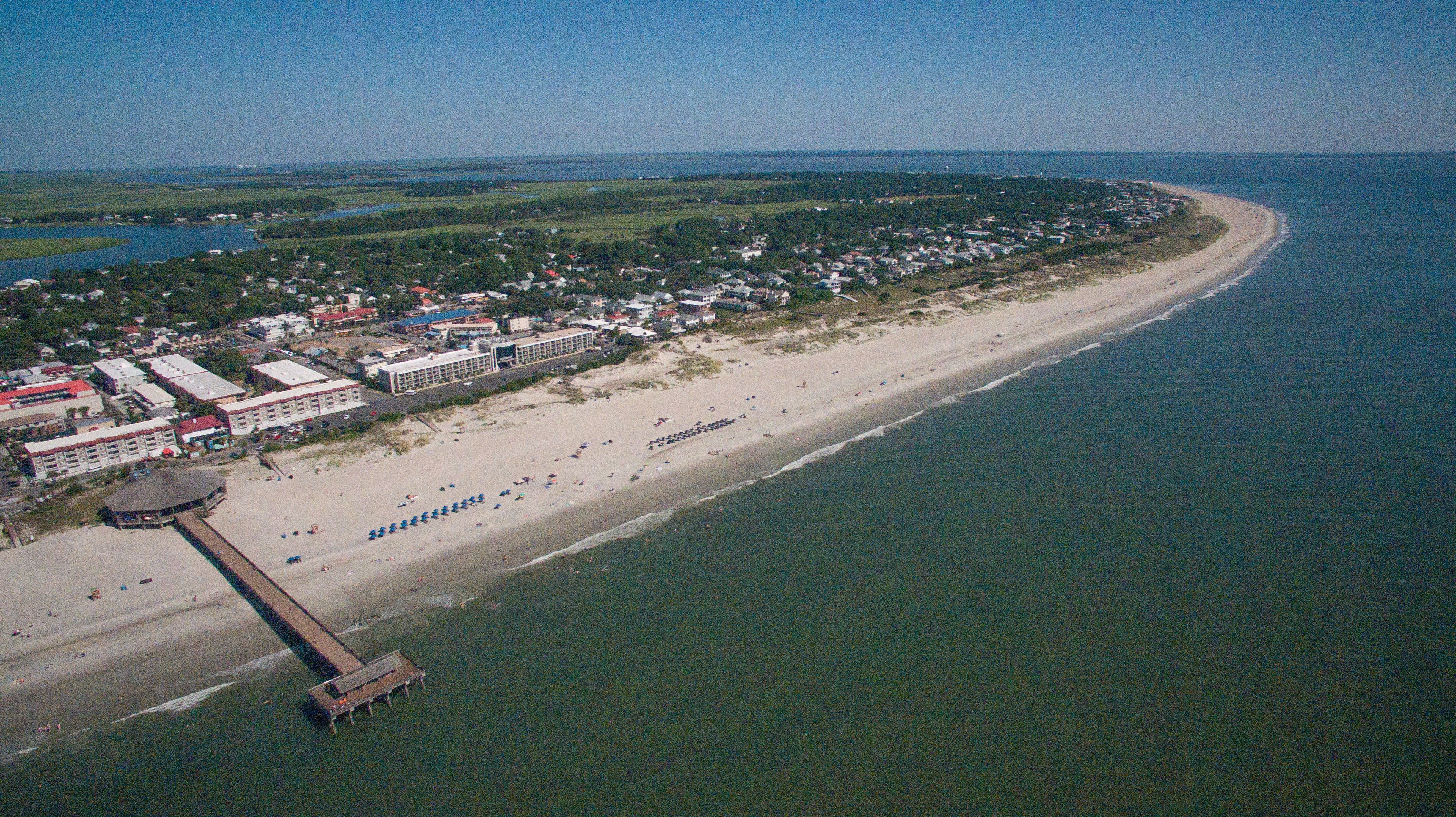 Luftaufnahme einer Küstenlandschaft mit Strand, einem Steg und einer Reihe von Gebäuden entlang des Ufers.
