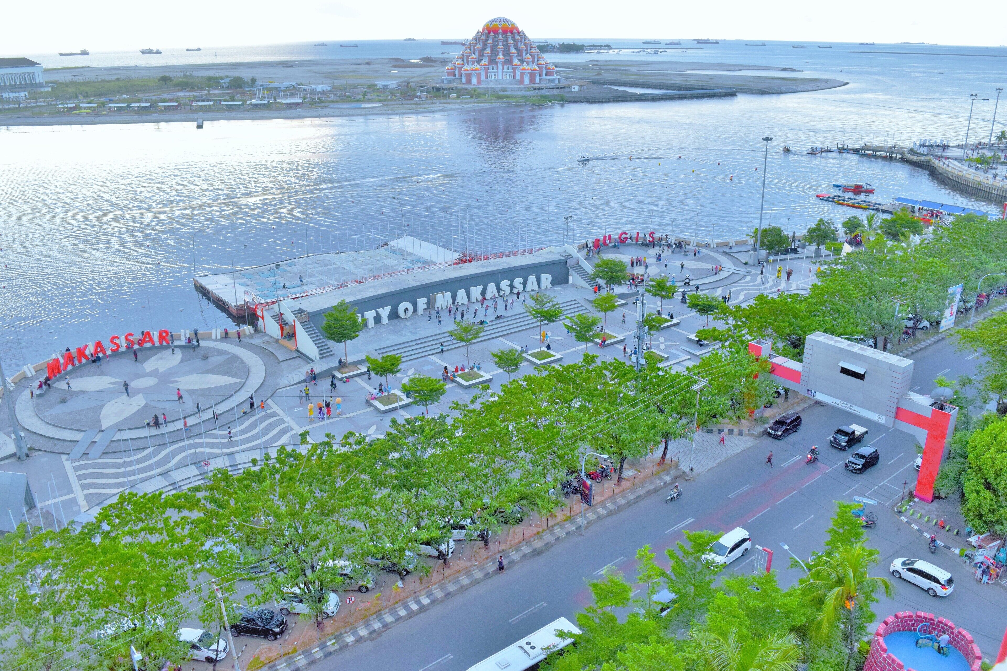 A waterfront area with a prominent building labeled "City of Makassar" and a large, colorful monument in the background.
