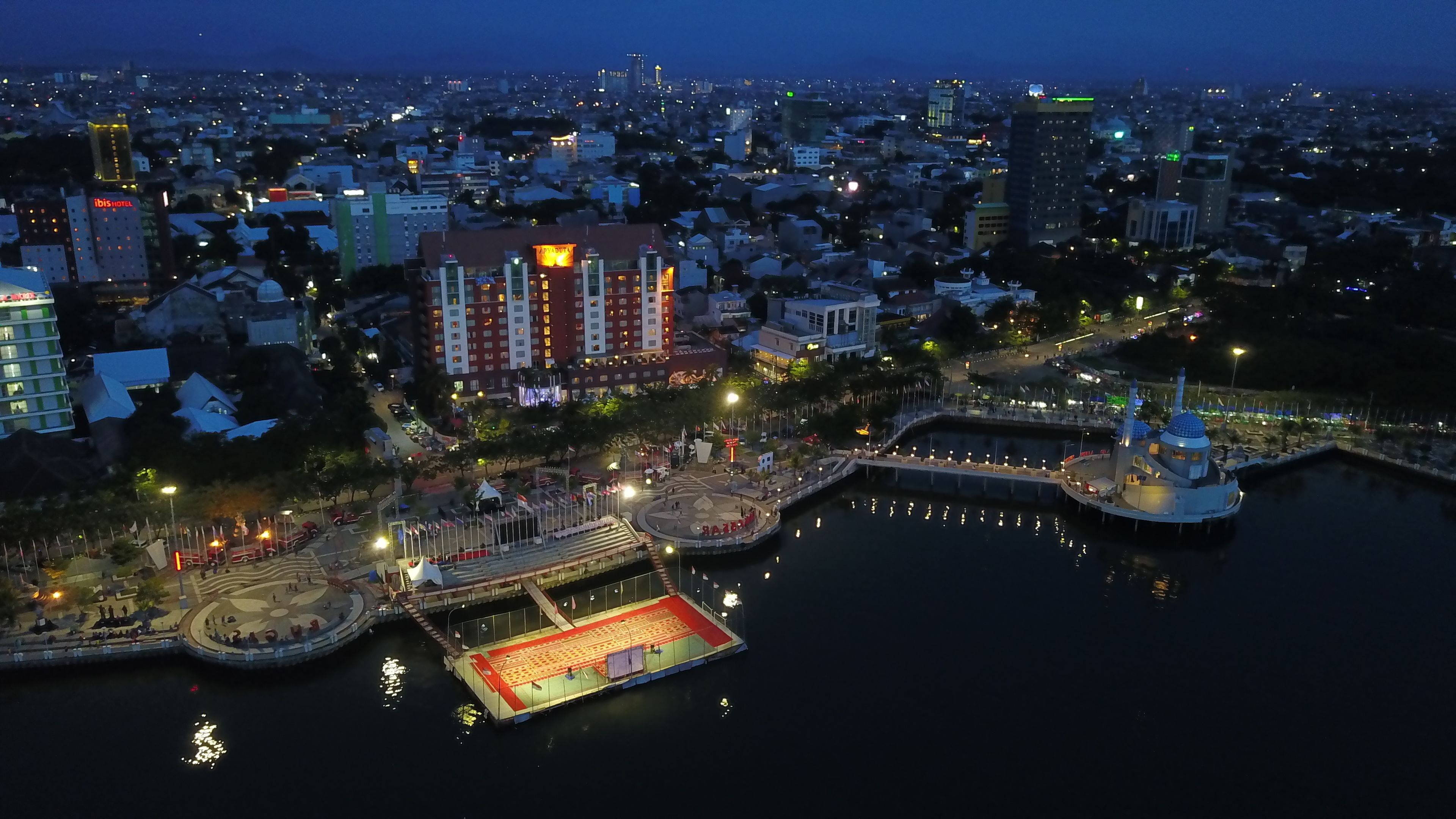 A waterfront area with a large open space, possibly for events, surrounded by buildings including a hotel with a red and white facade.
