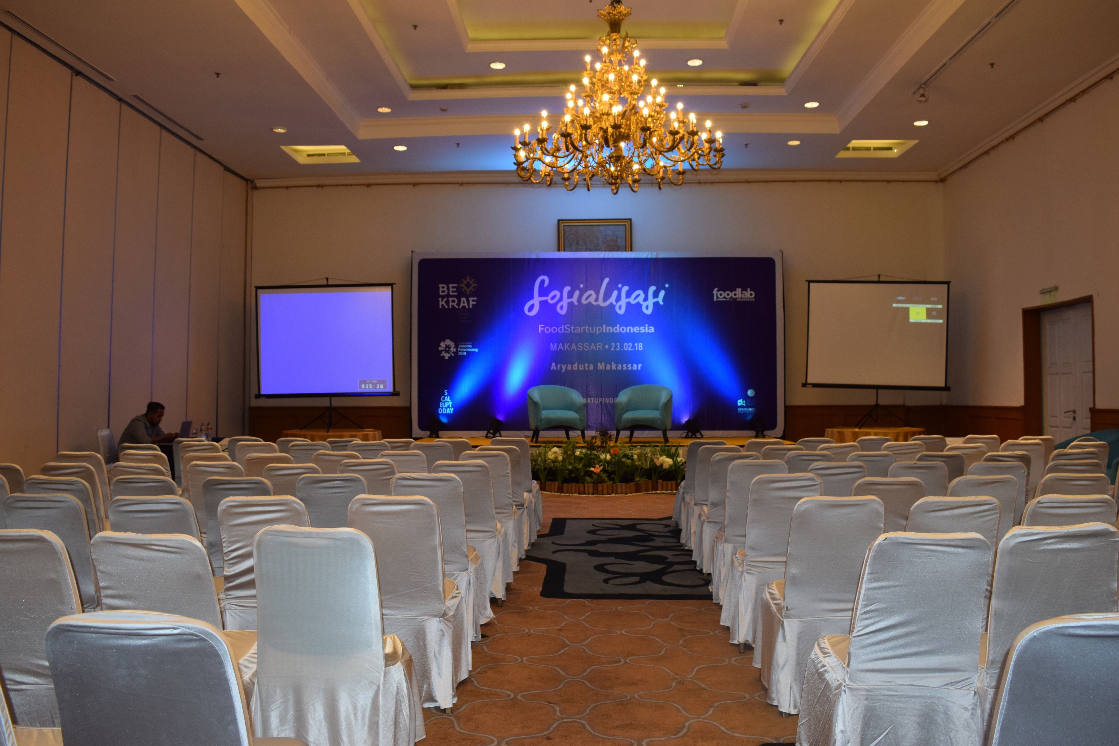 A conference room with rows of chairs, a stage with a presentation screen, and a chandelier.
