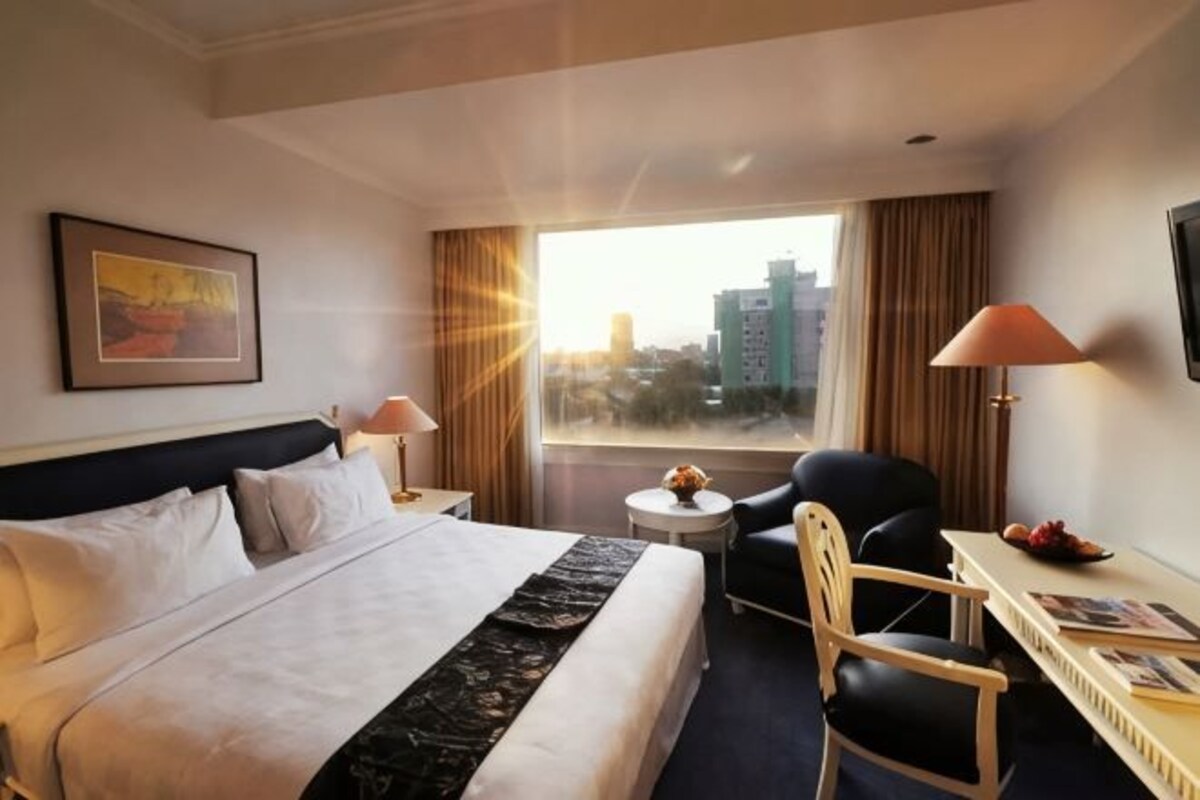 Habitación de hotel con cama, escritorio, silla, lámpara y vistas a la ciudad.