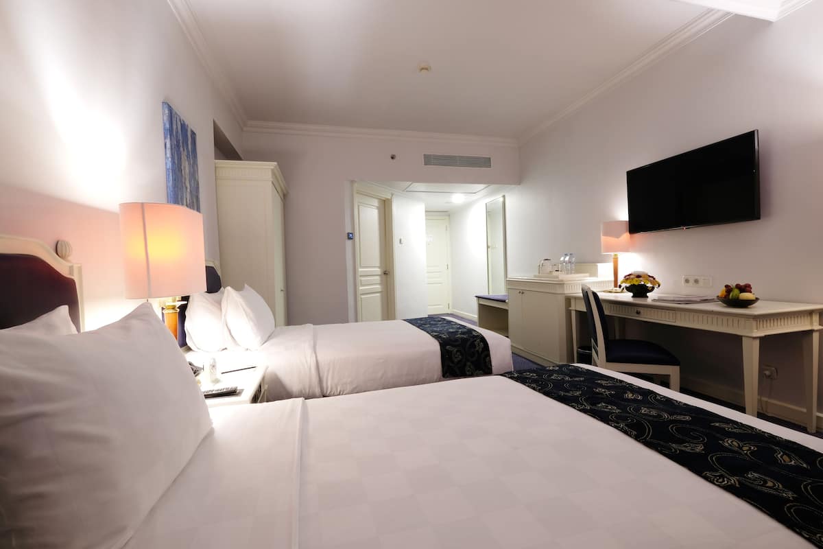 Habitación de hotel con dos camas, un escritorio, un televisor y un baño visible a través de una puerta abierta.