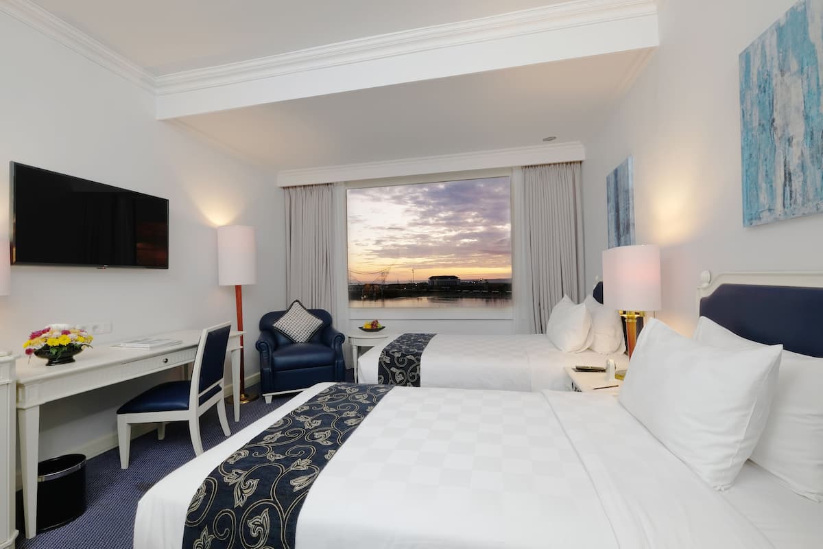 Habitación de hotel con dos camas, un escritorio, una silla, un televisor y vistas al atardecer sobre el agua.
