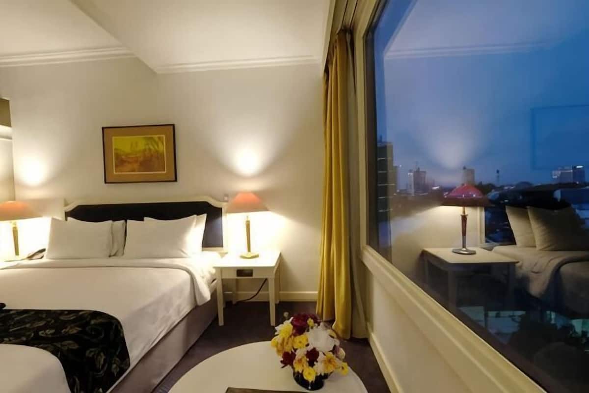 Habitación de hotel con cama, dos mesitas de noche, un florero y un ventanal con vistas a la ciudad.