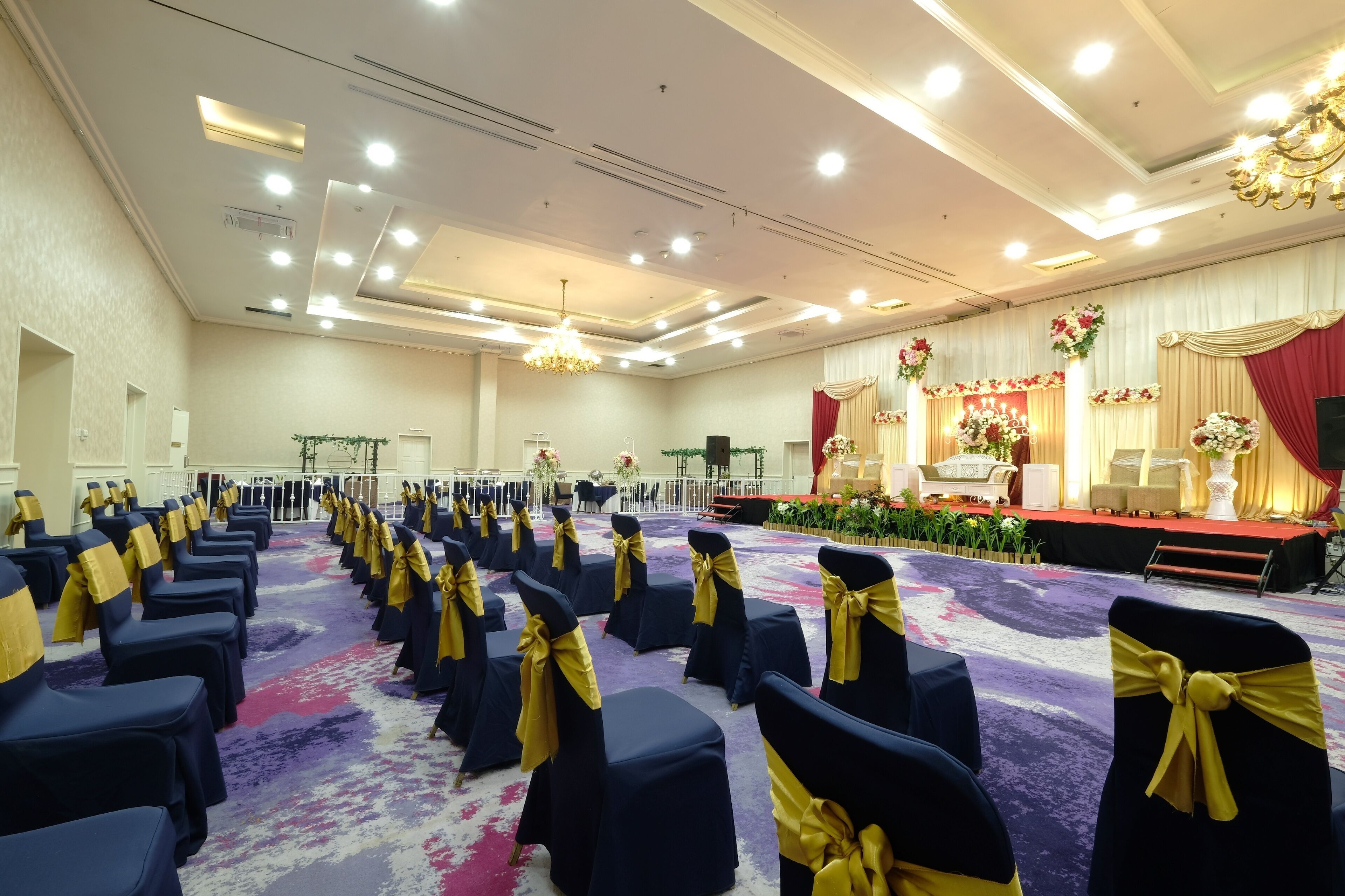 A banquet hall with rows of tables and chairs, a stage with floral decorations, and a chandelier.