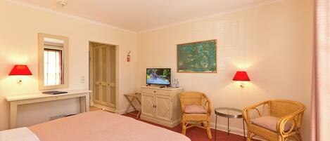 Habitación de hotel con cama, televisor sobre un mueble, dos sillones de mimbre, una mesita y dos lámparas de pared.