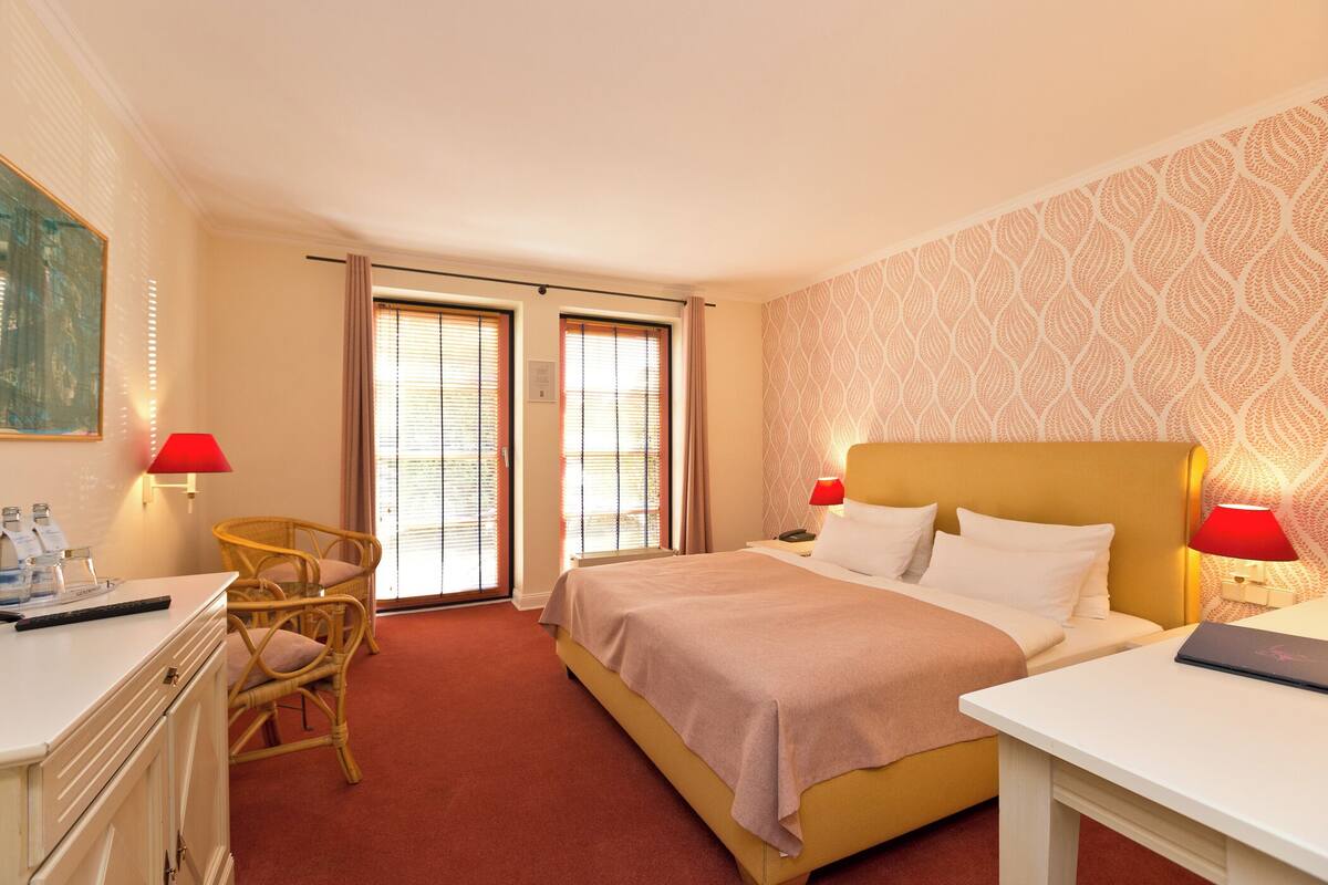 A hotel room with a bed, two bedside tables, a desk, a chair, and a window with blinds.