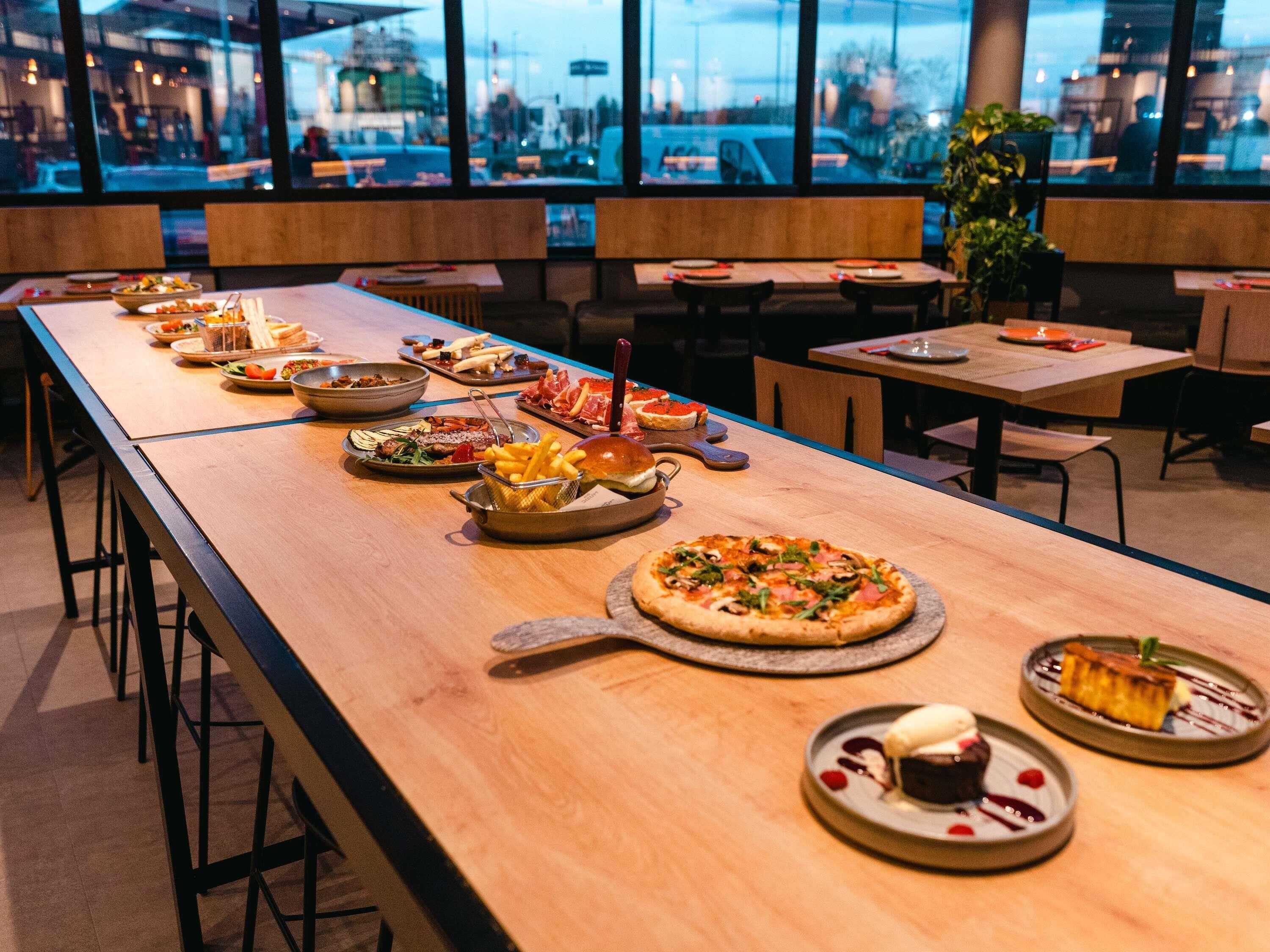 A long dining table with various dishes, including pizza and desserts, in a modern restaurant setting with large windows.