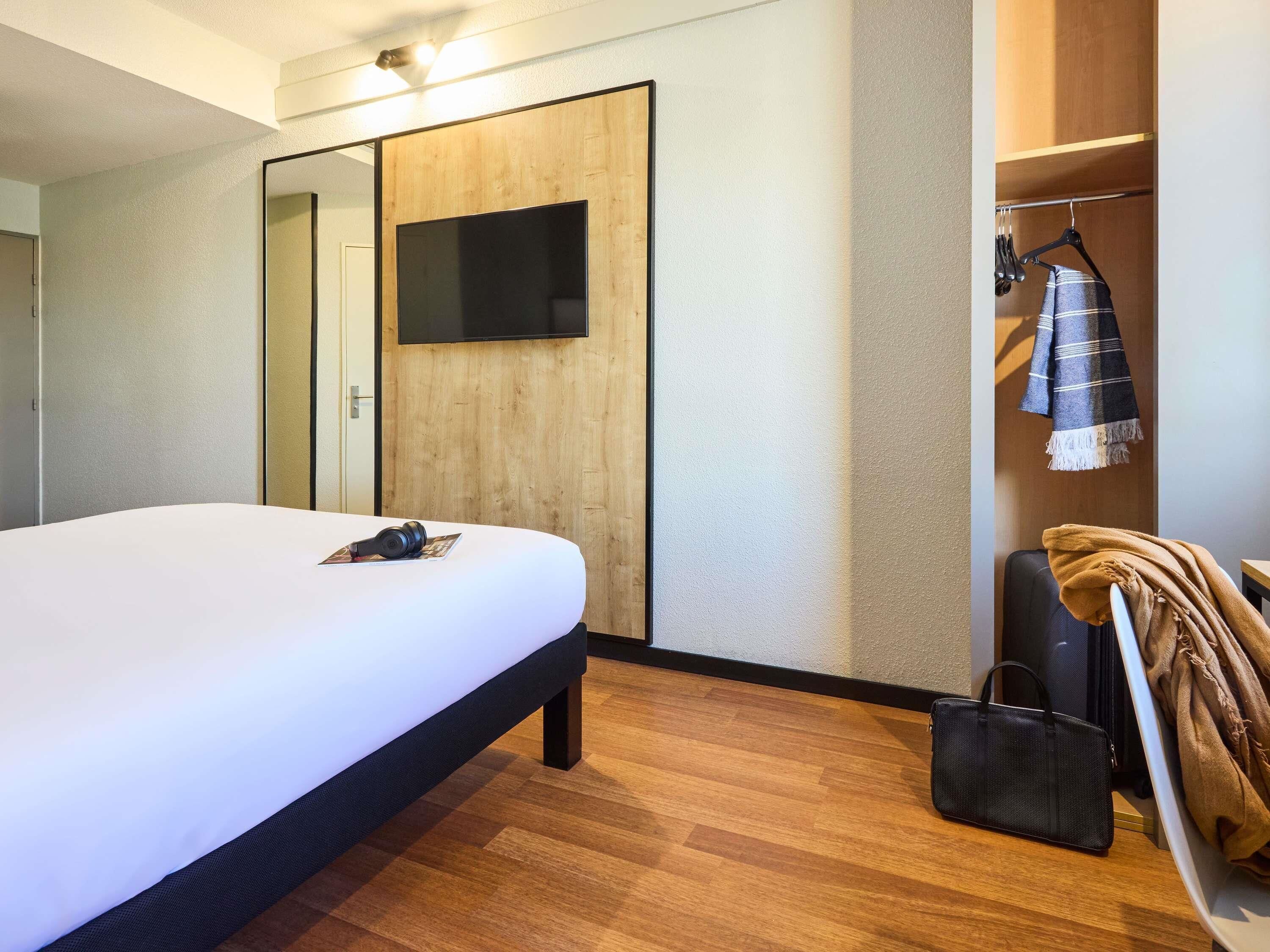 A hotel room with a bed, a TV mounted on a wooden wall, a wardrobe, and a coat rack with a blanket.