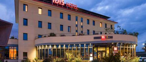 A modern hotel with a prominent 'ibis Hotel' sign, a glass facade, and a landscaped entrance area.