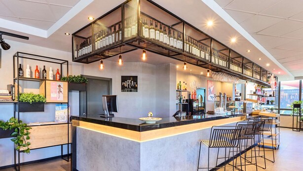 A modern bar area with a central counter, bar stools, and a mezzanine shelf displaying bottles. - ibis Valladolid (Valladolid)