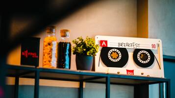 Une cassette audio étiquetée « Breakfast 90 » est posée sur une étagère avec divers objets, notamment une bouteille, une plante en pot et une boîte.