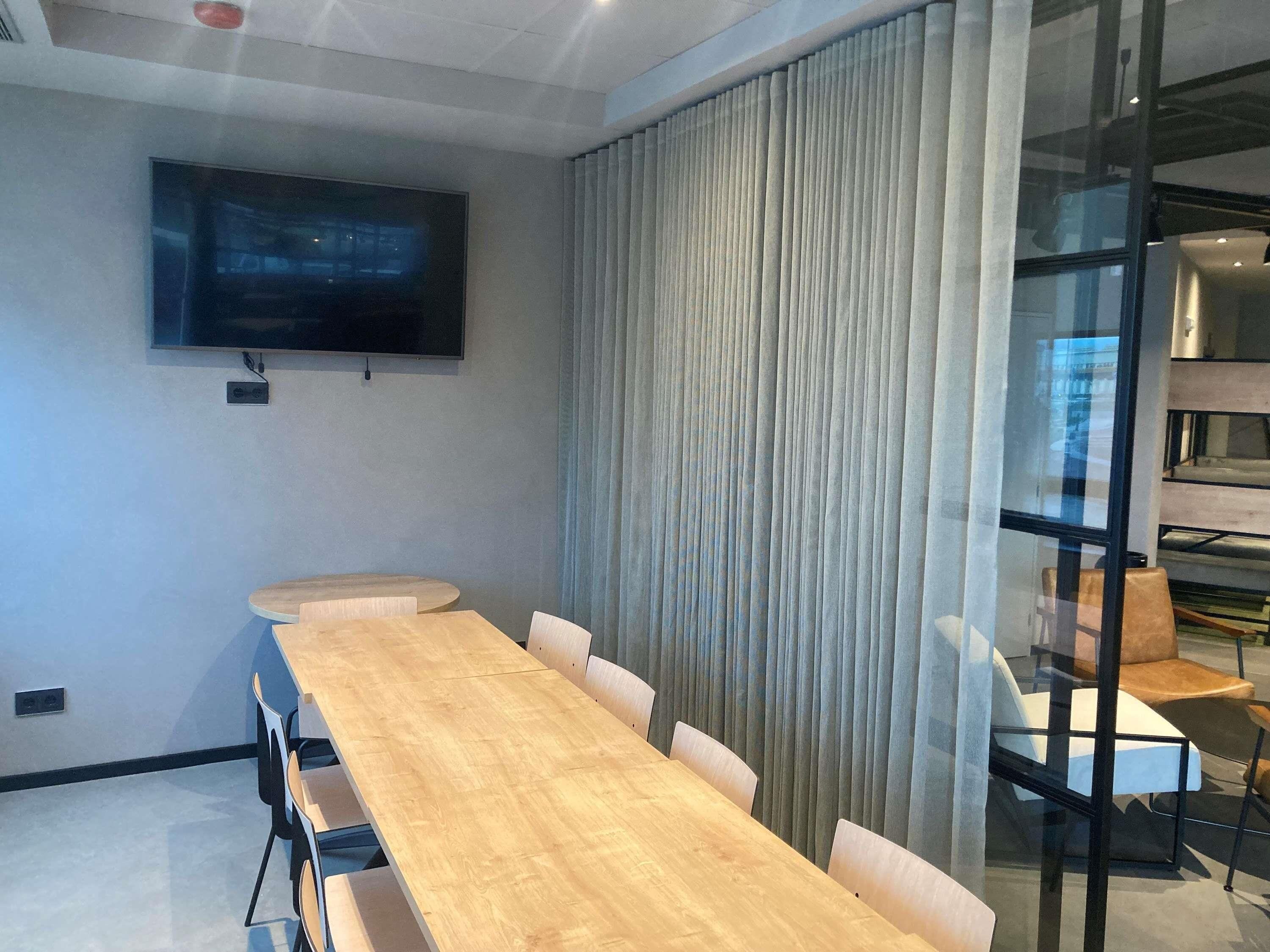 A conference room with a long wooden table, six chairs, a flat-screen TV mounted on the wall, and a large window with curtains.