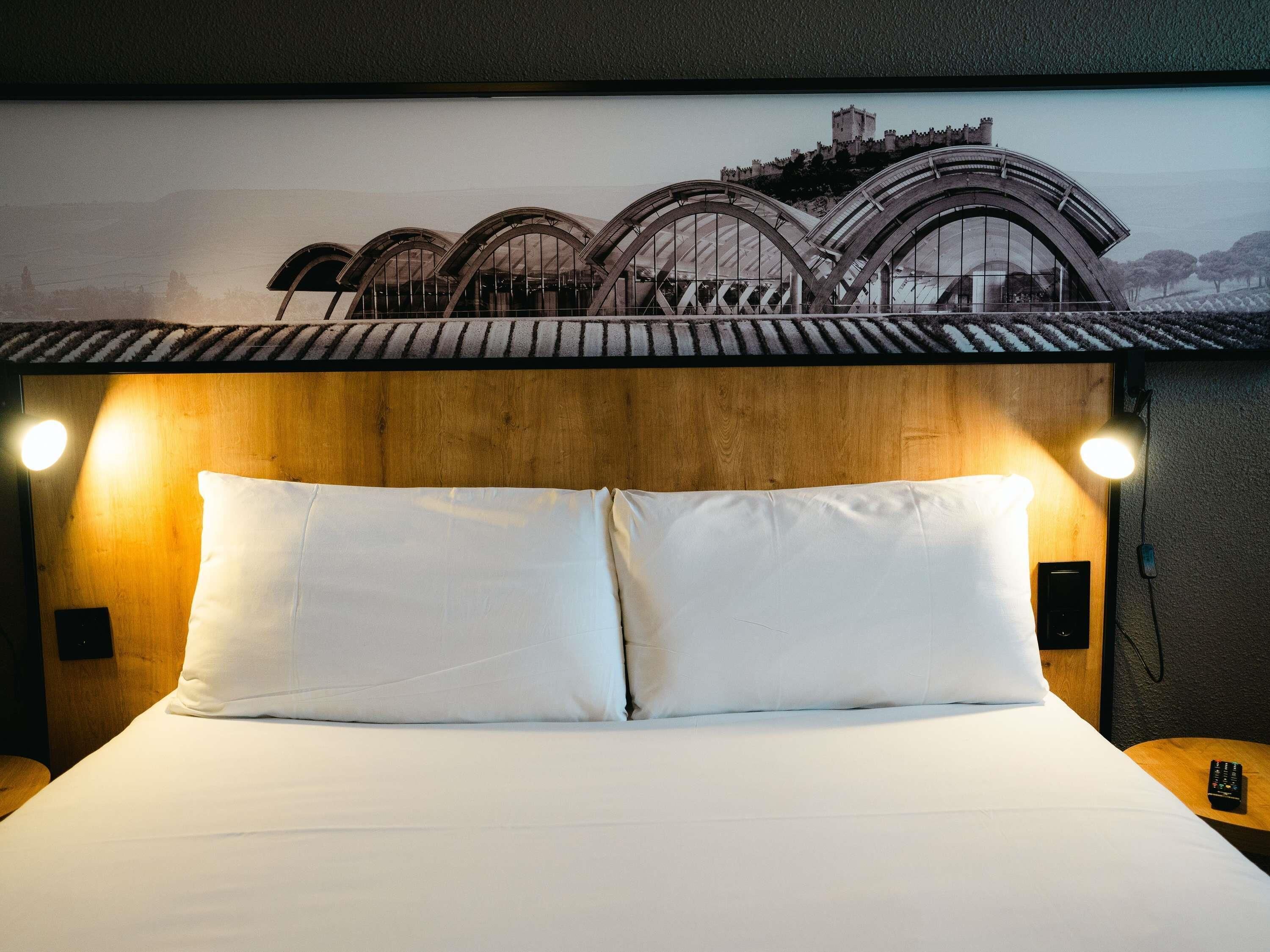 Een bed met witte lakens, twee bedlampen en een tv aan de muur.