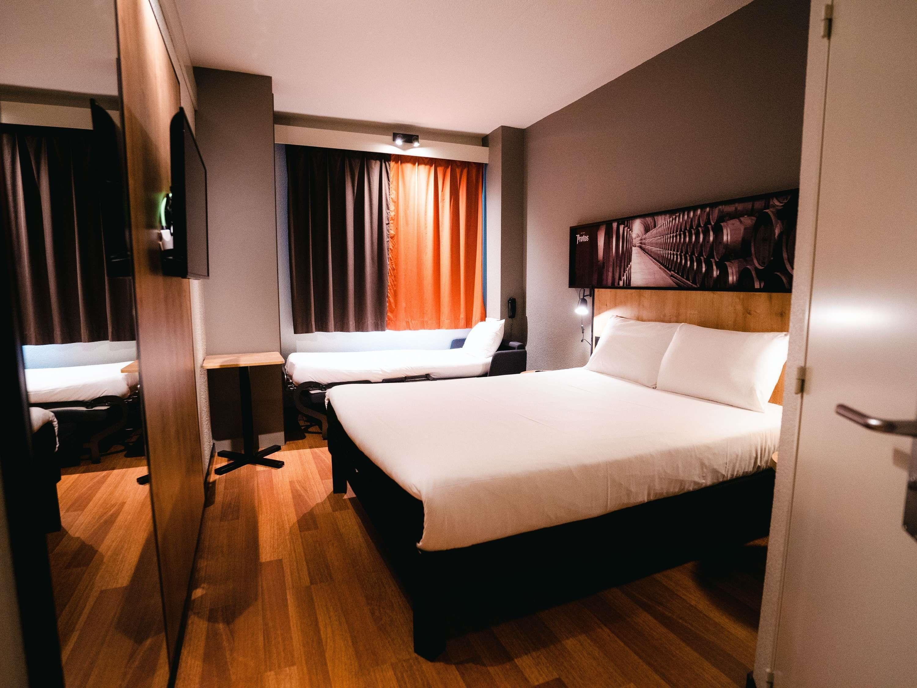 A hotel room with a large bed, a desk, a chair, a TV, and a window with curtains.