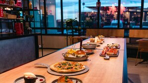 A long table with various dishes, including pizzas and desserts, in a modern restaurant setting with large windows. - ibis Valladolid (Valladolid)