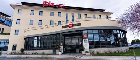 A modern hotel with a curved glass facade, a prominent red logo, and a sign indicating it is part of the ibis hotel chain.