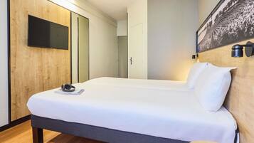 A hotel room with a large bed, a flat-screen TV mounted on the wall, a bedside table with a tablet, and a wooden headboard.