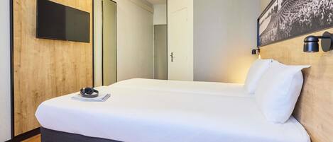 A hotel room with a large bed, a flat-screen TV mounted on the wall, a bedside table with a tablet, and a wooden headboard.