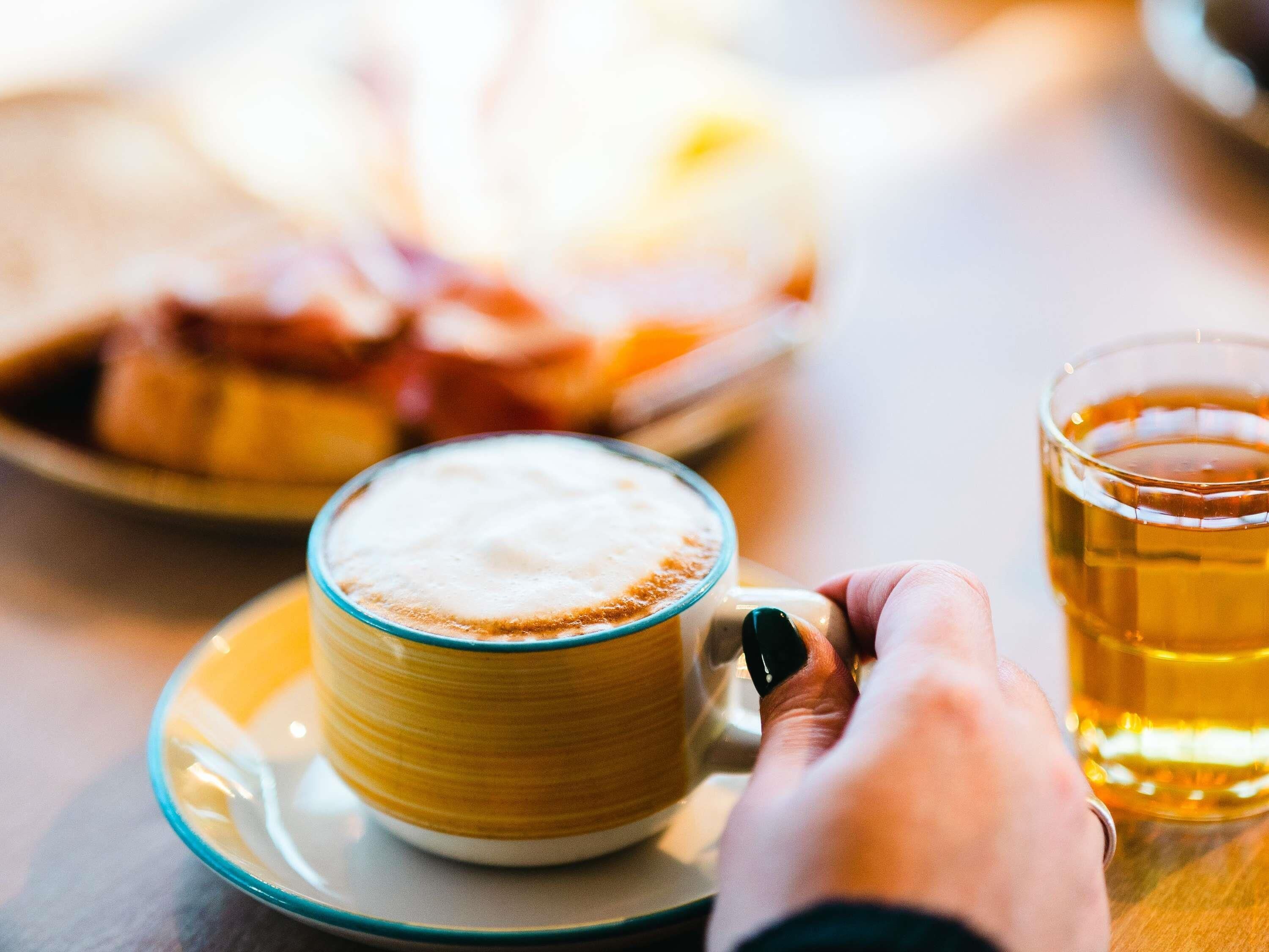 A hand holding a mug of coffee with a saucer, a glass of beer, and a blurred plate of food in the background.