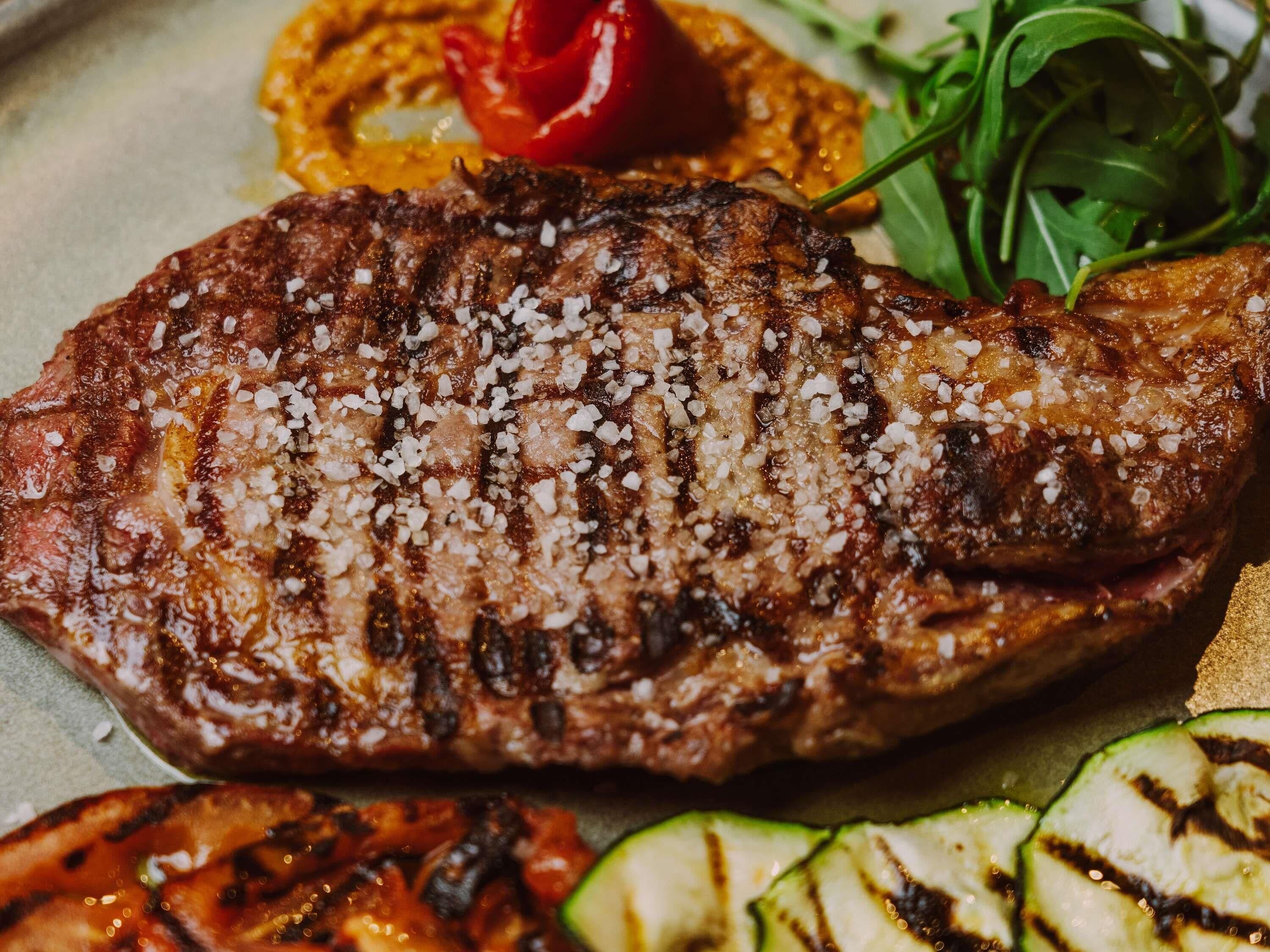 A grilled steak with salt on top, accompanied by grilled vegetables and a red pepper.