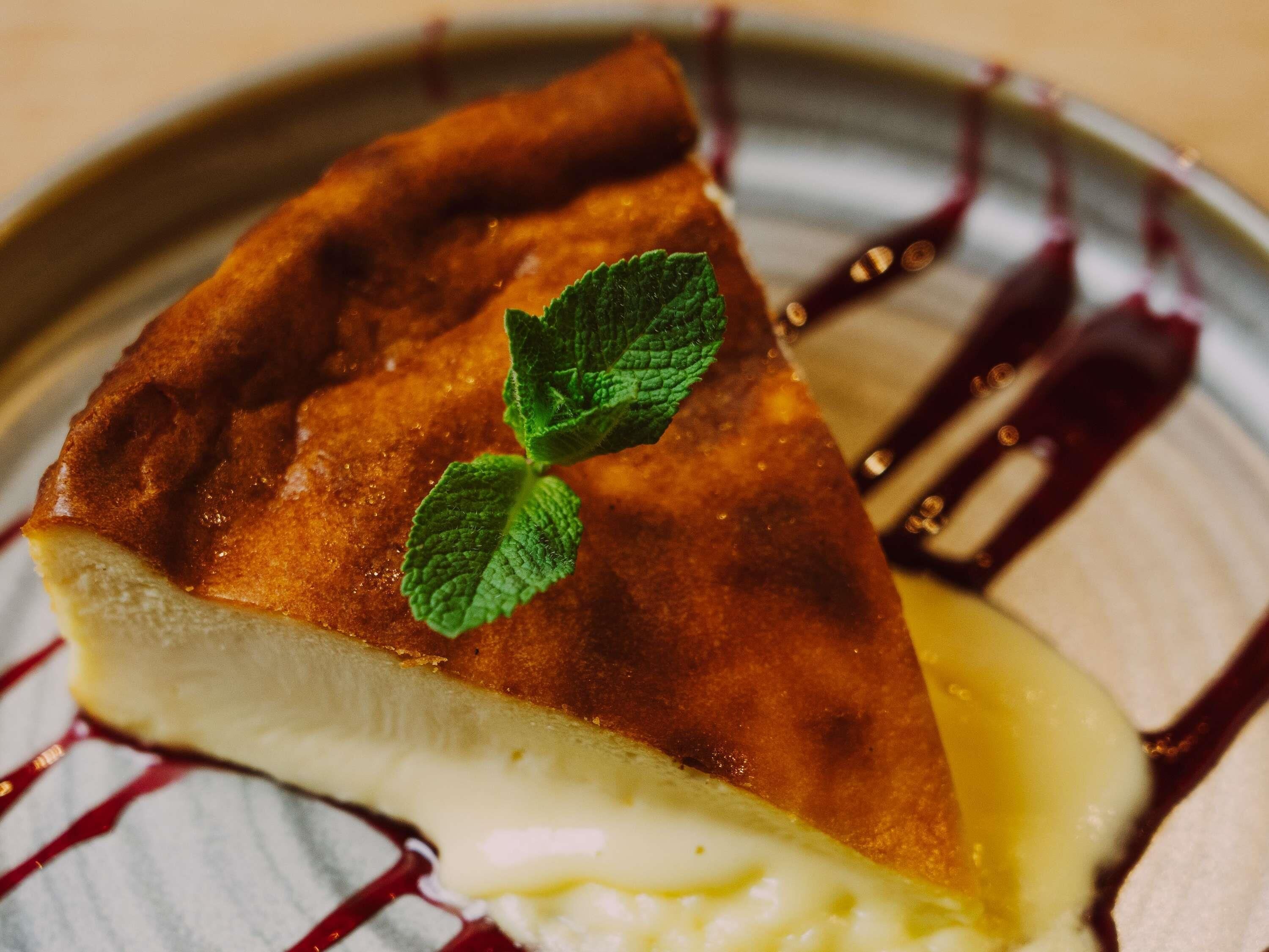 Una porción de pastel de queso con corteza dorada, una hoja de menta como adorno y un hilo de salsa roja sobre un plato.