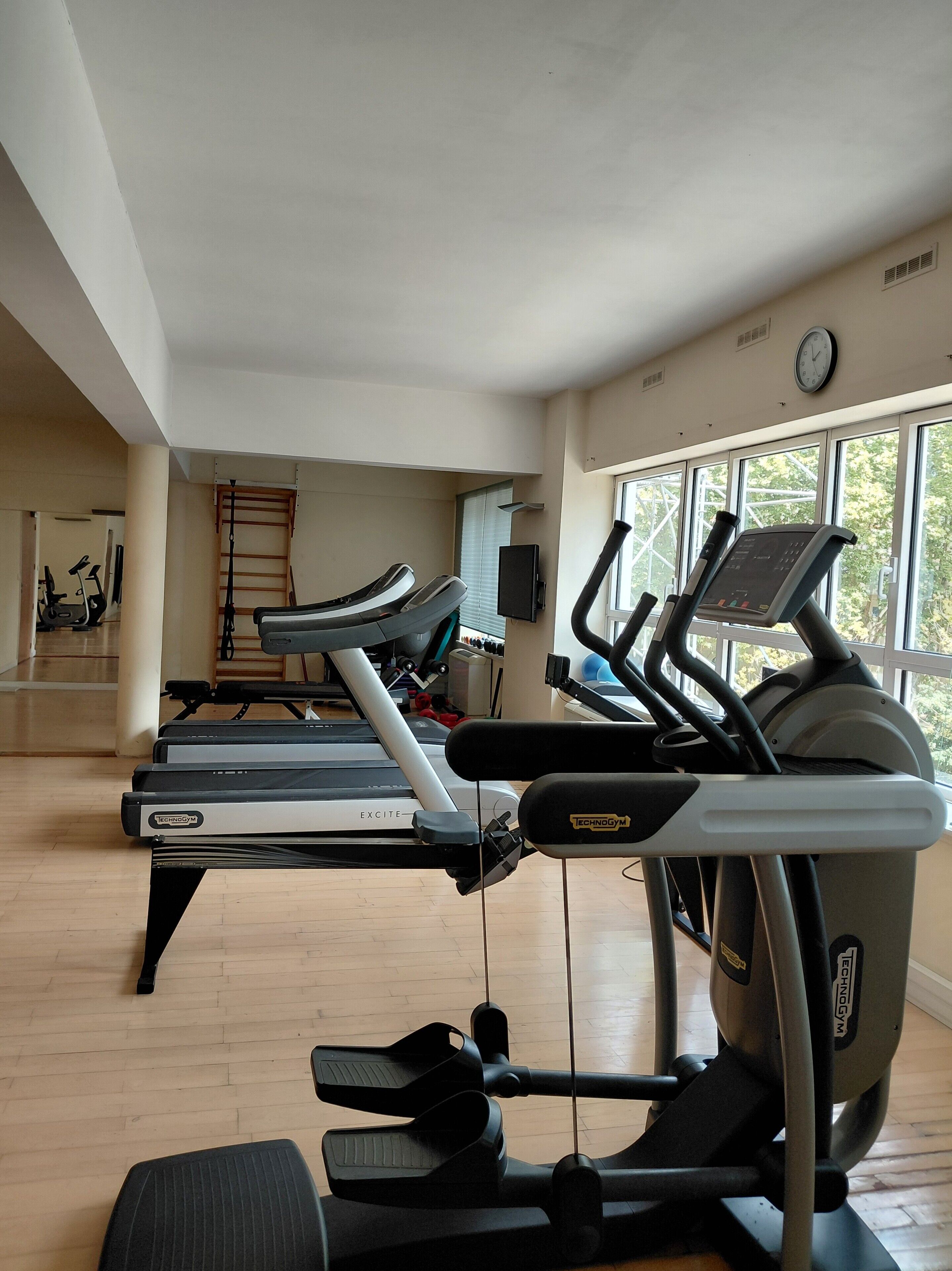 Une salle de sport équipée de tapis de course, d’un vélo d’appartement et d’un rameur. Il y a une fenêtre donnant sur des arbres et une horloge au mur.