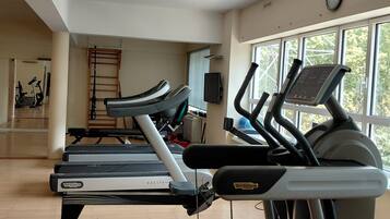 Une salle de sport équipée de tapis de course, d’un vélo d’appartement et d’un rameur. Il y a une fenêtre donnant sur des arbres et une horloge au mur.