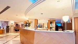 A modern hotel lobby with a reception desk, a large vase of flowers, and a digital screen displaying a scenic view.