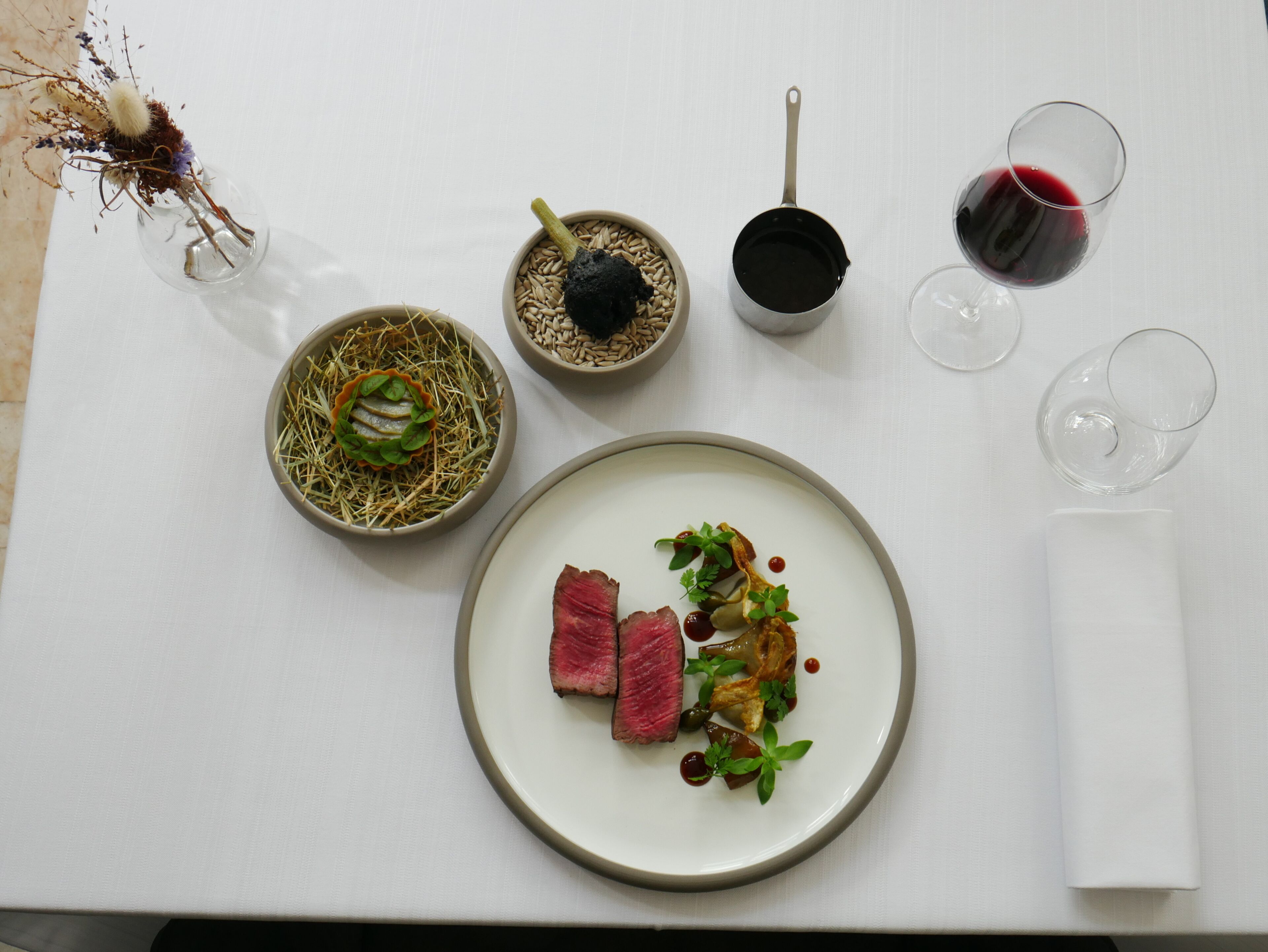 A dining table with a plate of food, a bowl of grains, a small pot, and two glasses of red wine.