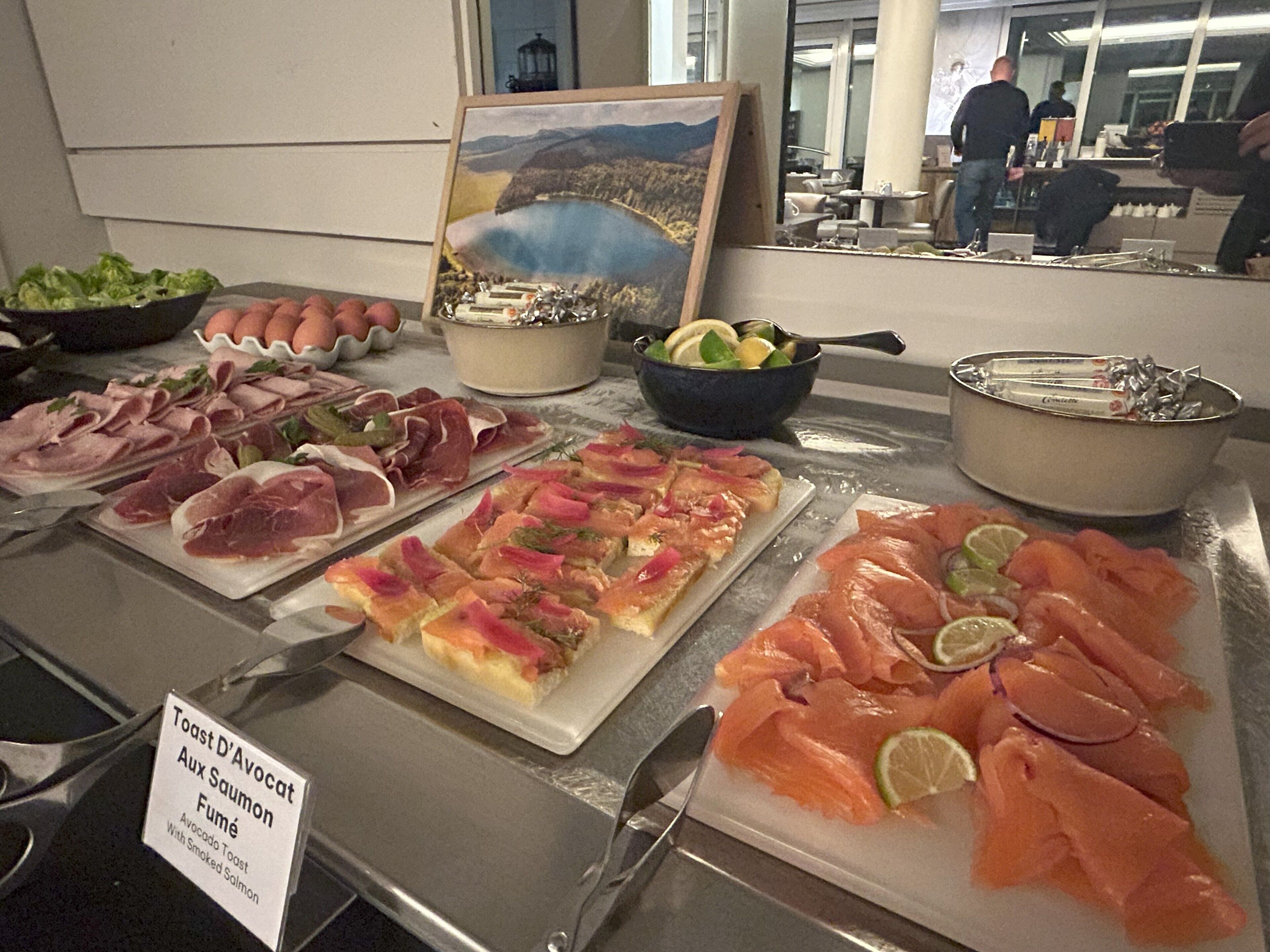 En buffet med forskellige spiser, heriblandt røget laks, skinke og en ret med 'Toast D'Avocat Aux Saumon Fumé'.