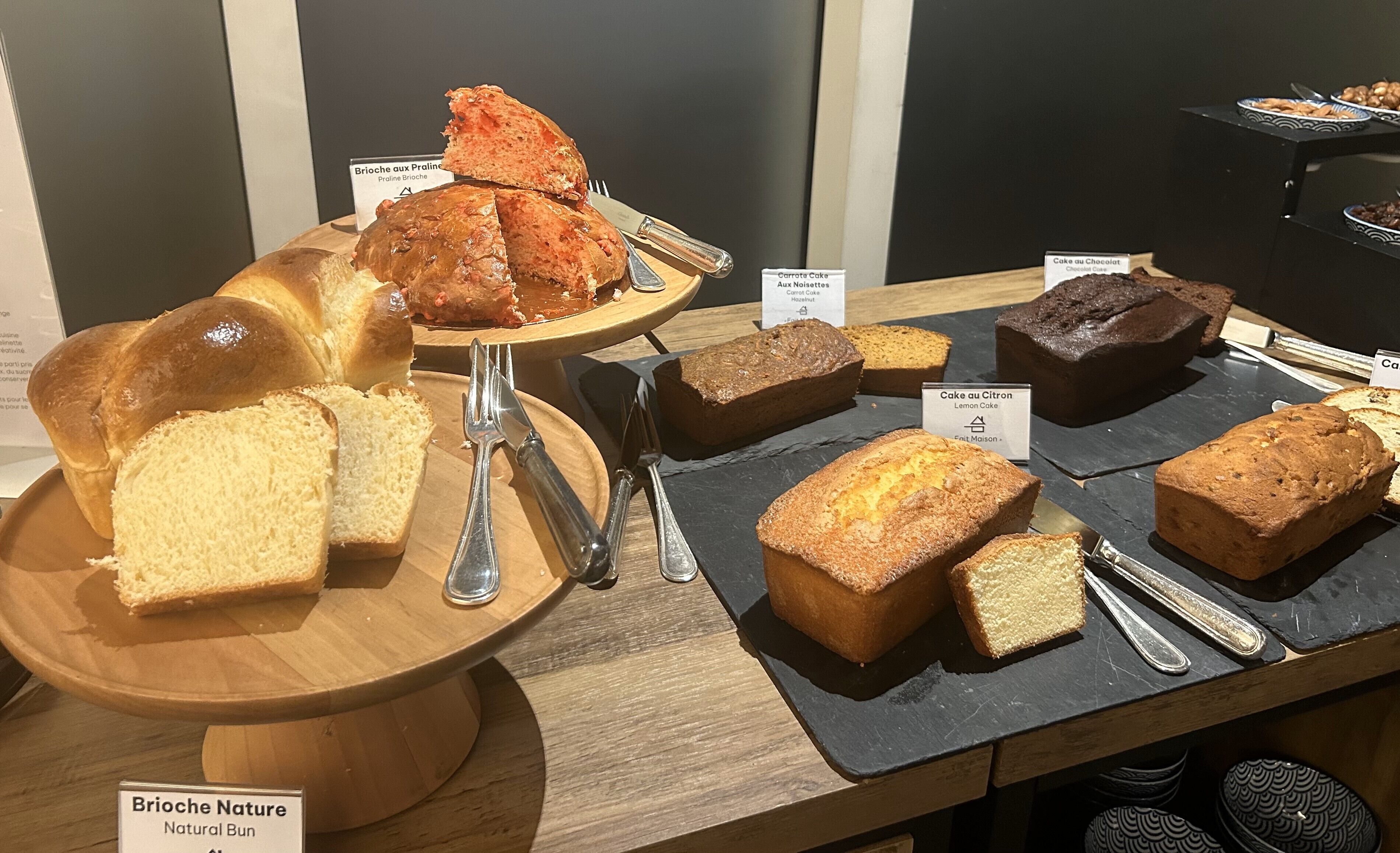 Diverse broden en gebak uitgestald, met labels, waaronder Brioche Nature, worteltaart en citroentaart.