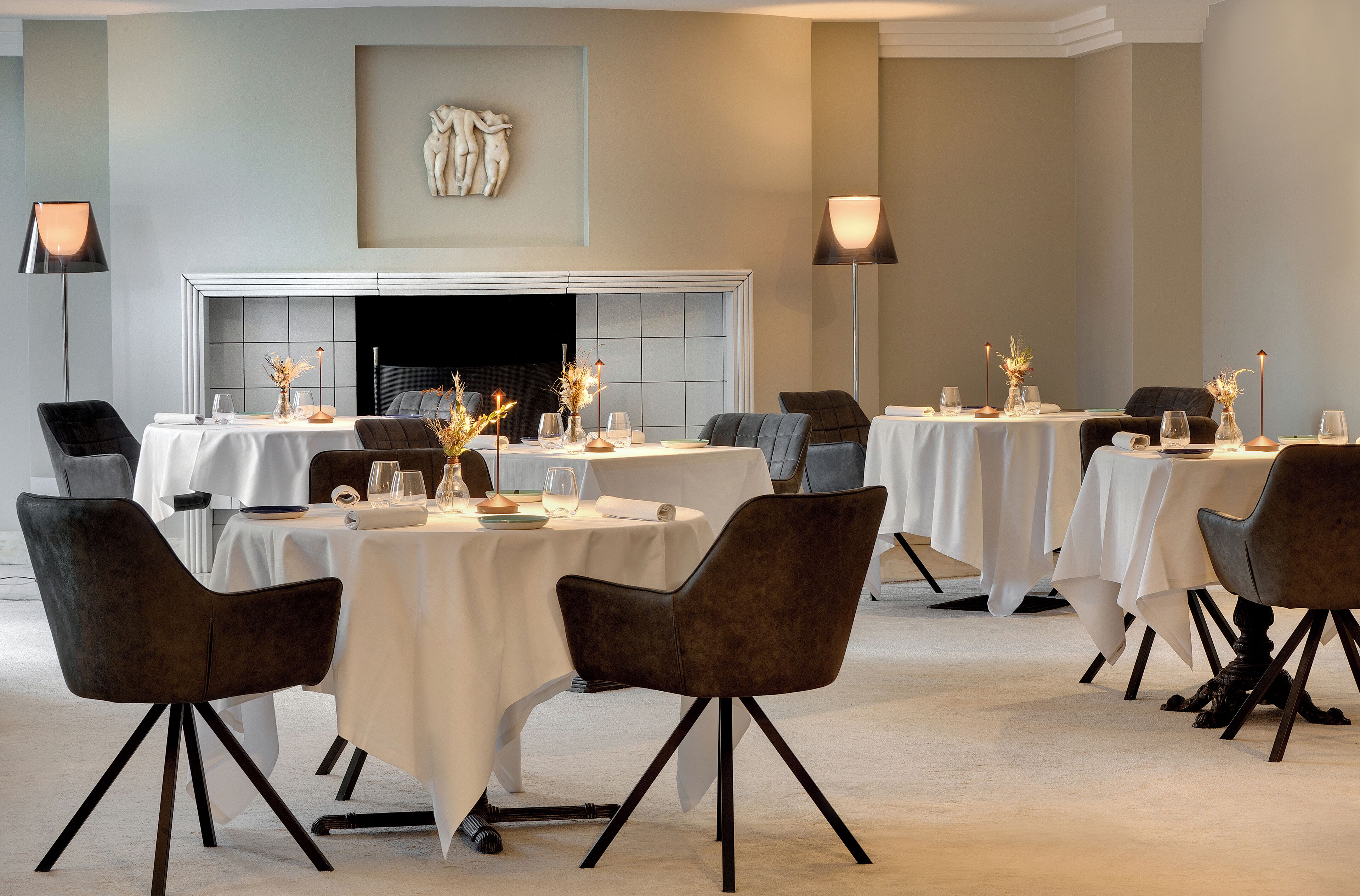Une salle à manger avec plusieurs tables rondes, chacune recouverte d’une nappe blanche et entourée de chaises foncées. Deux grandes lampes sur pied sont placées de part et d’autre de la pièce, et une cheminée est visible au fond.