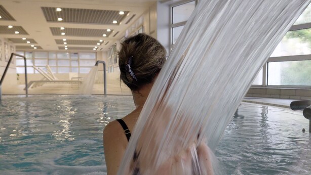 A person in a hot tub with a waterfall feature, indoor pool, and large windows.