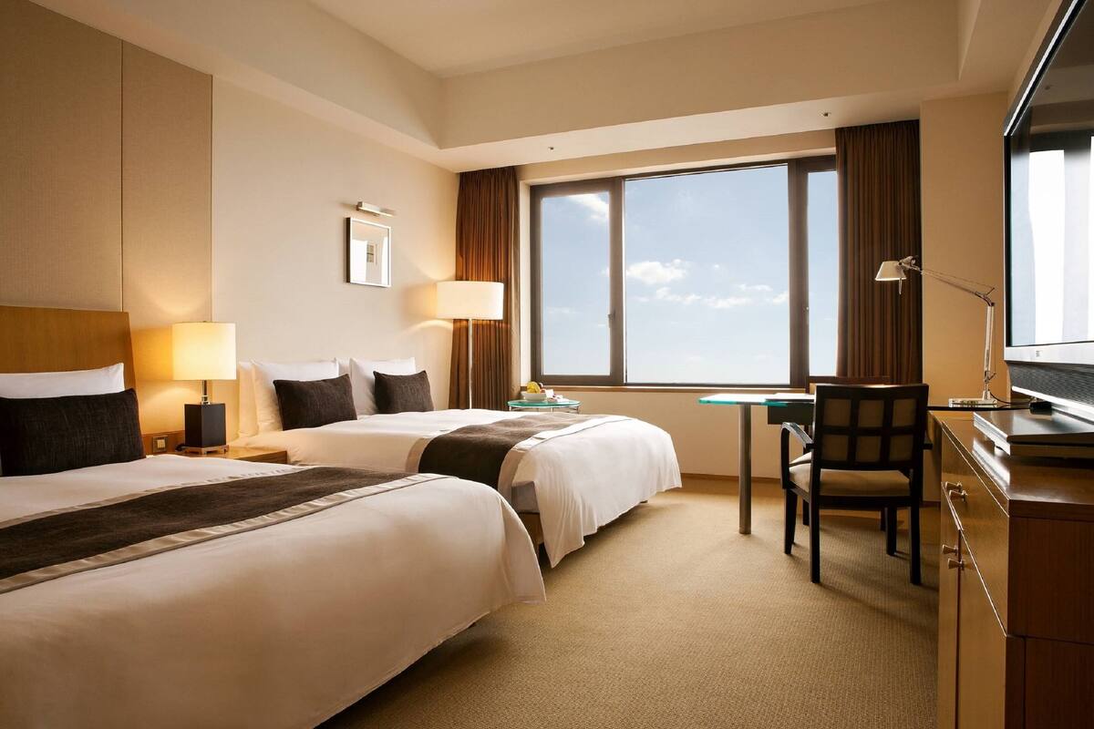 A hotel room with two beds, a desk, a chair, a TV, and a large window with a view of the sky.