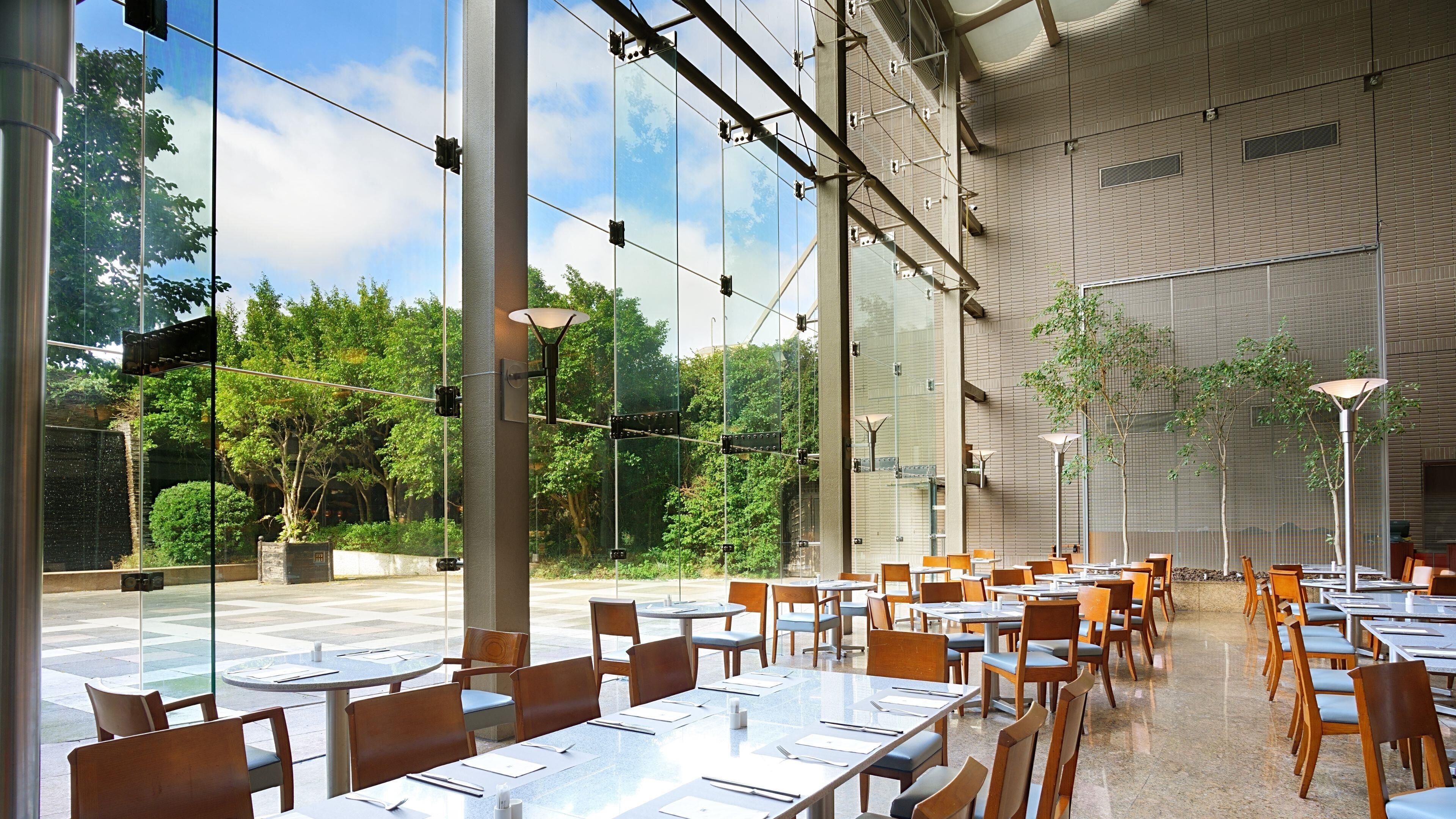 A spacious indoor dining area with large windows, modern furniture, and a view of greenery outside.