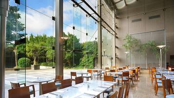 A spacious indoor dining area with large windows, modern furniture, and a view of greenery outside.