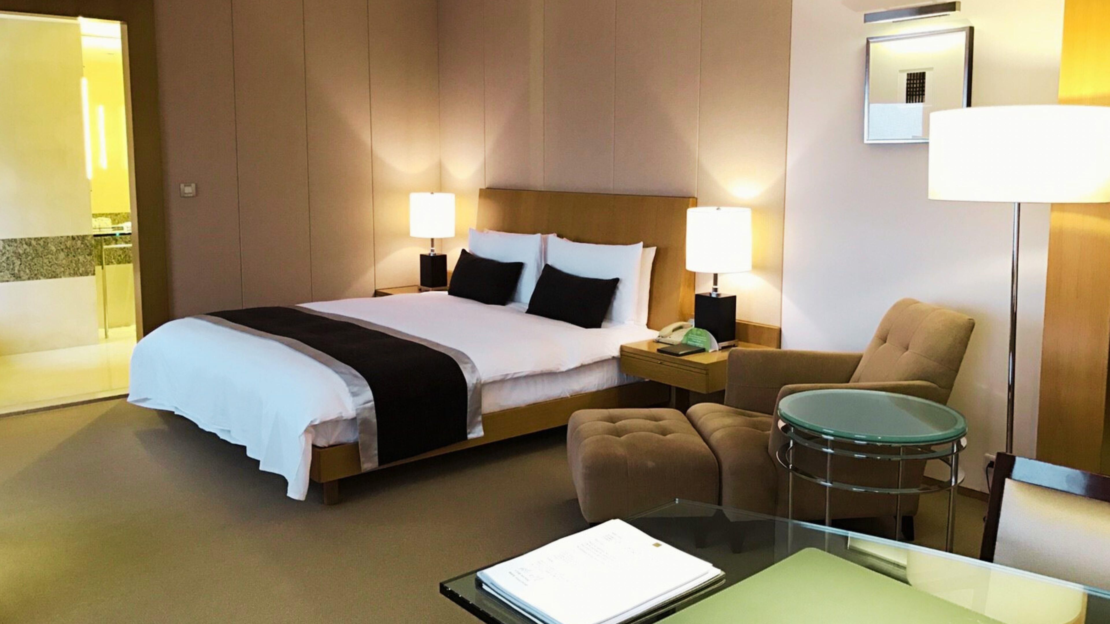 A hotel room with a bed, bedside lamps, a chair, a small table, and a desk with a lamp.