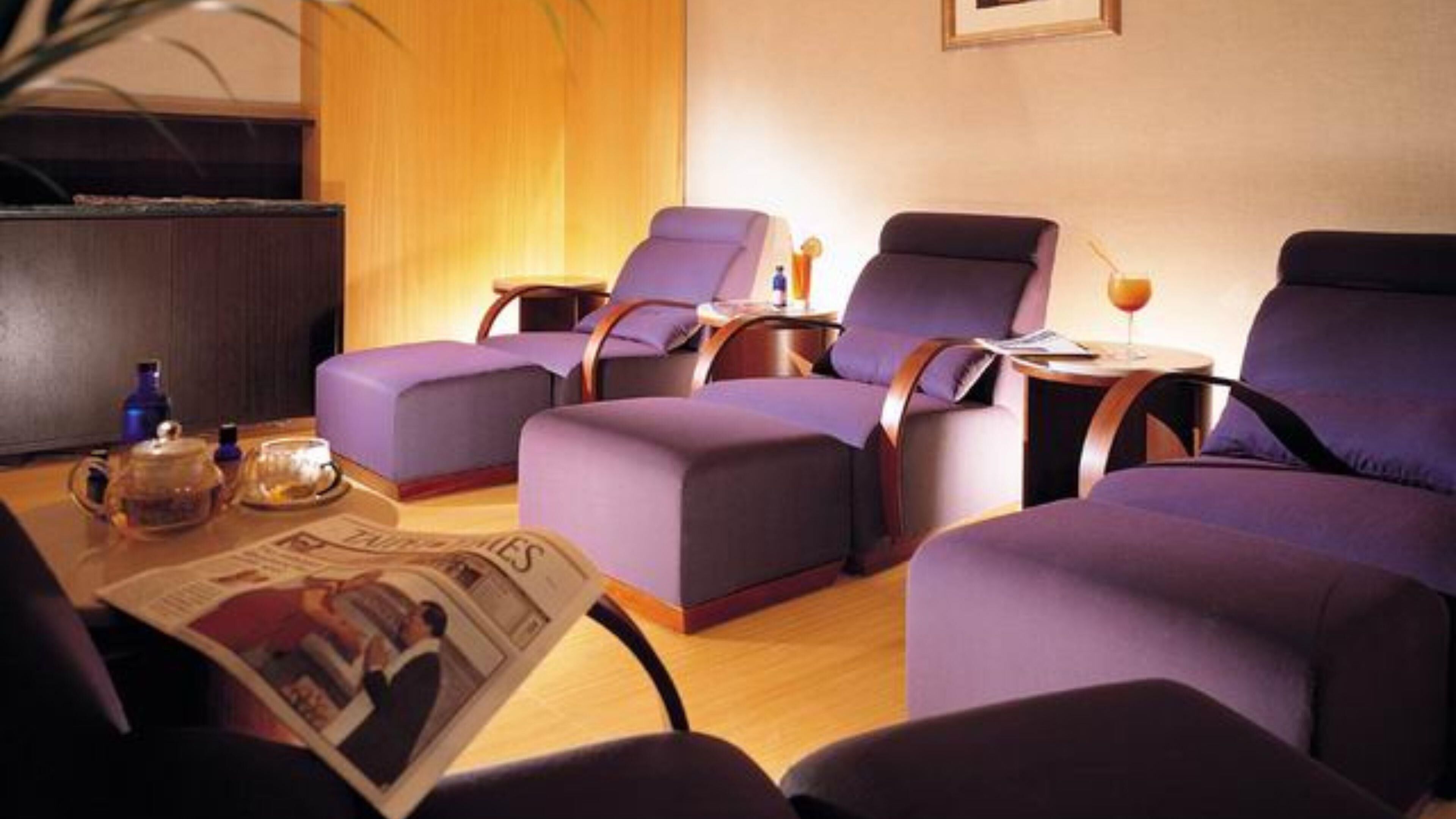 A room with four purple armchairs arranged around a wooden coffee table, a small side table with a lamp, and a television on the left.