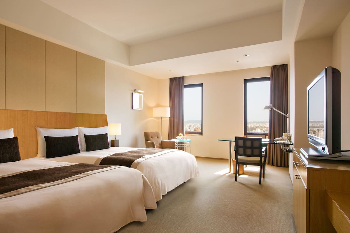 A hotel room with two beds, a desk, a chair, a TV, and a city view.