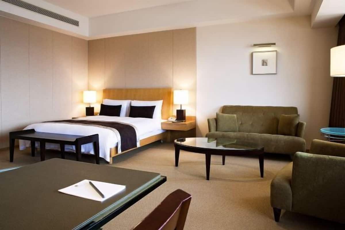A hotel room with a bed, two bedside tables with lamps, a sofa, a coffee table, and a desk with a chair.