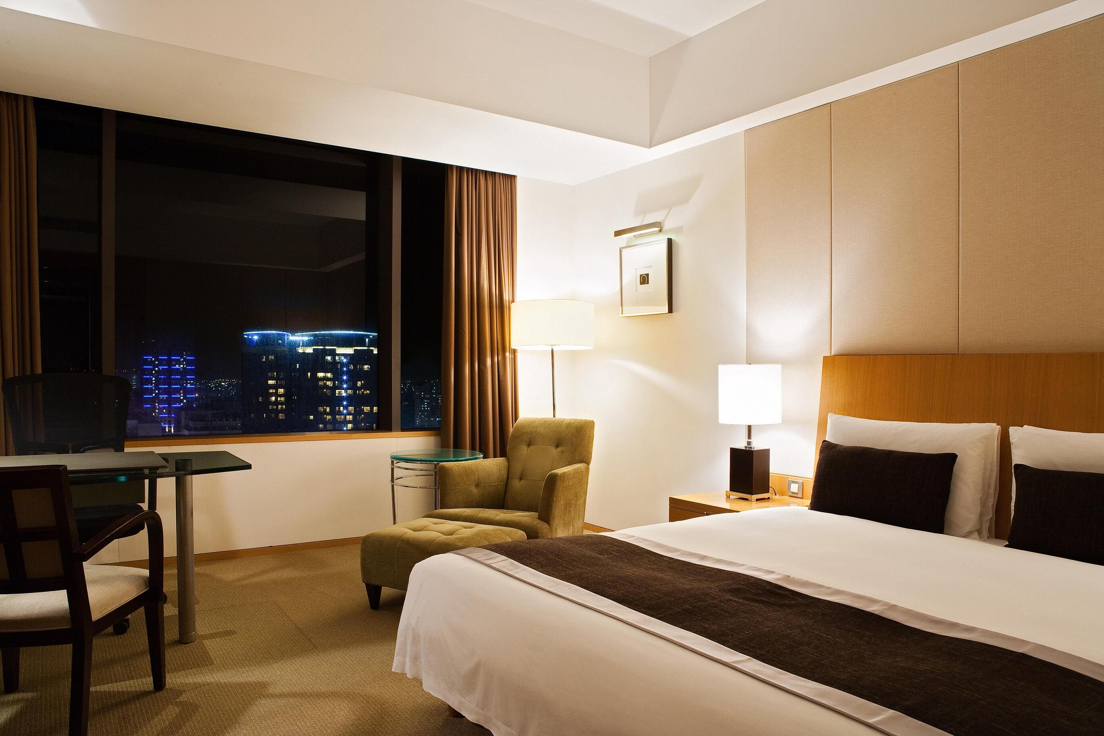 Ein Hotelzimmer mit Bett, Schreibtisch, Stuhl, Lampe und Blick auf die nächtliche Stadt.
